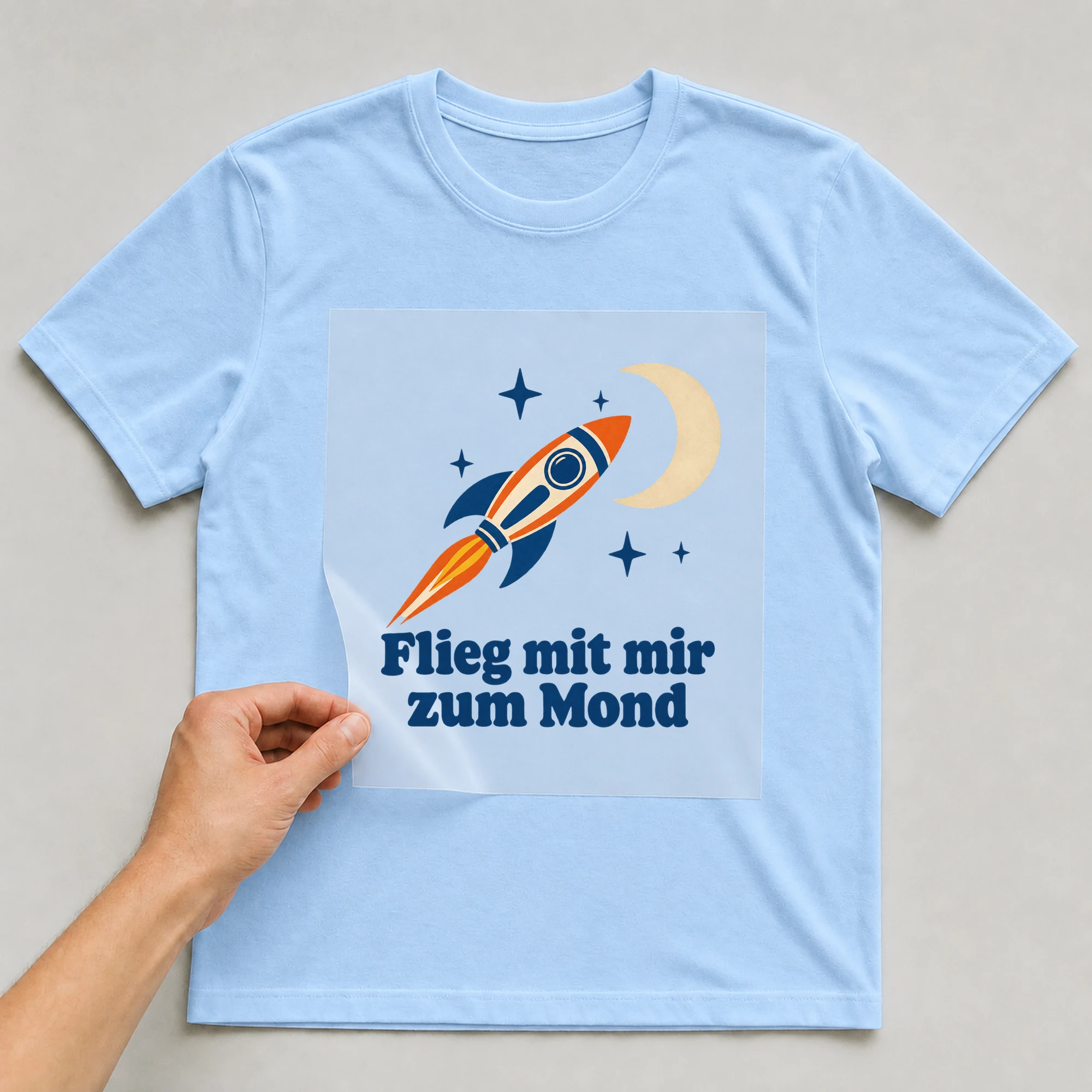 Bügelbild „Rakete und Mond mit Schrift“ auf babyblauem T-Shirt, Folie wird aufgelegt
