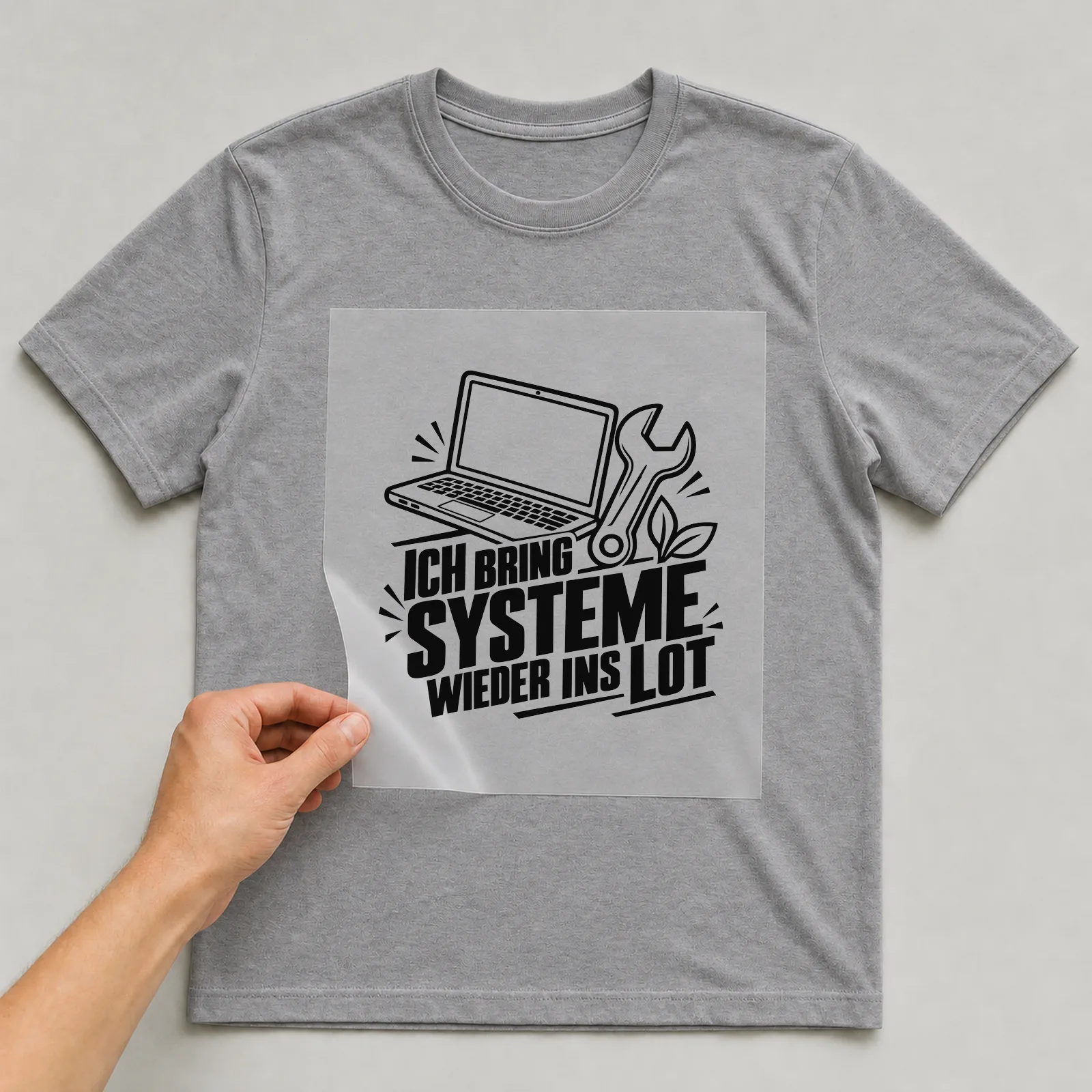 Bügelbild „Ich bring Systeme wieder ins Lot“ auf grauem T-Shirt, Folie wird aufgelegt