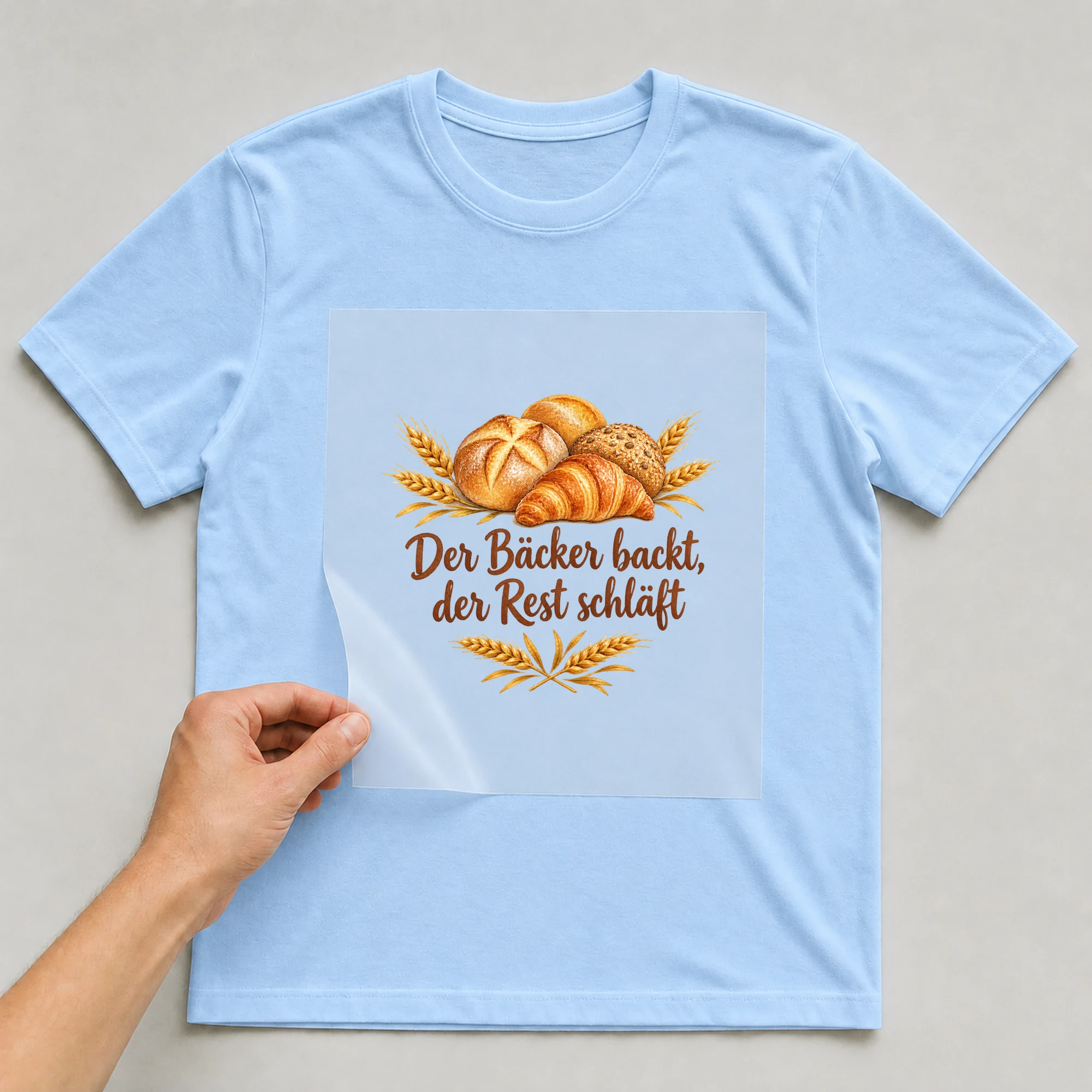 Bügelbild „Der Bäcker backt, der Rest schläft“ auf babyblauem T-Shirt, Folie wird aufgelegt