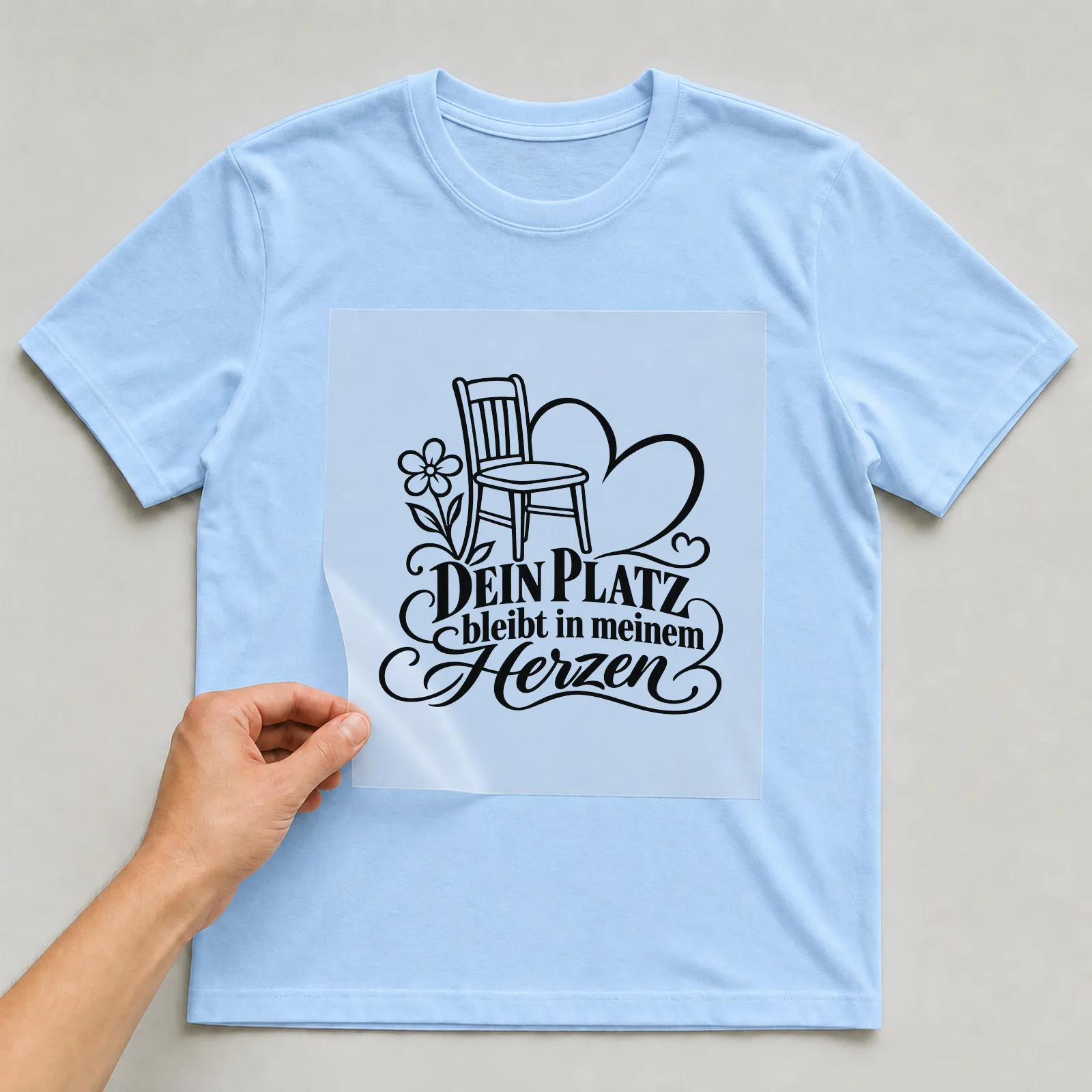 Bügelbild „Dein Platz bleibt in meinem Herzen“ auf babyblauem T-Shirt, Folie wird aufgelegt