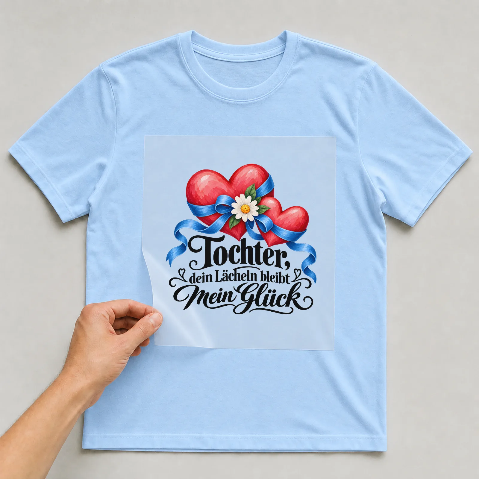 Bügelbild „Tochter, dein Lächeln bleibt mein Glück“ auf hellblauem T-Shirt, Folie wird aufgelegt