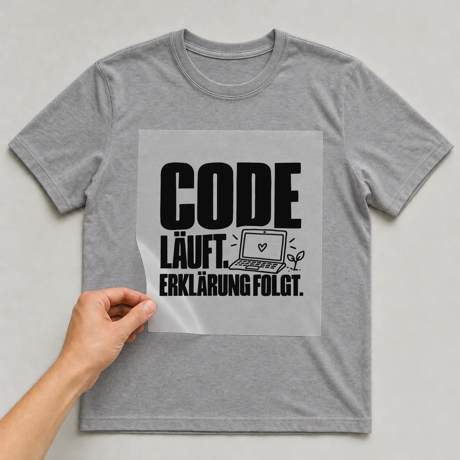 Bügelbild „Code läuft. Erklärung folgt.“ auf grauem T-Shirt, Folie wird aufgelegt
