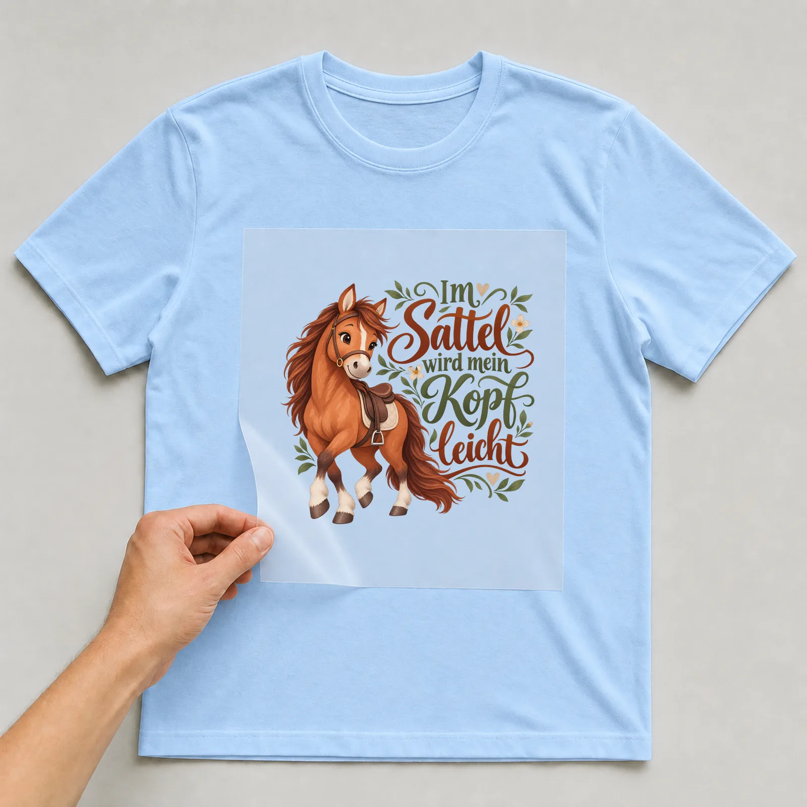Bügelbild „Im Sattel wird mein Kopf leicht“ auf hellblauem T-Shirt, Folie wird aufgelegt