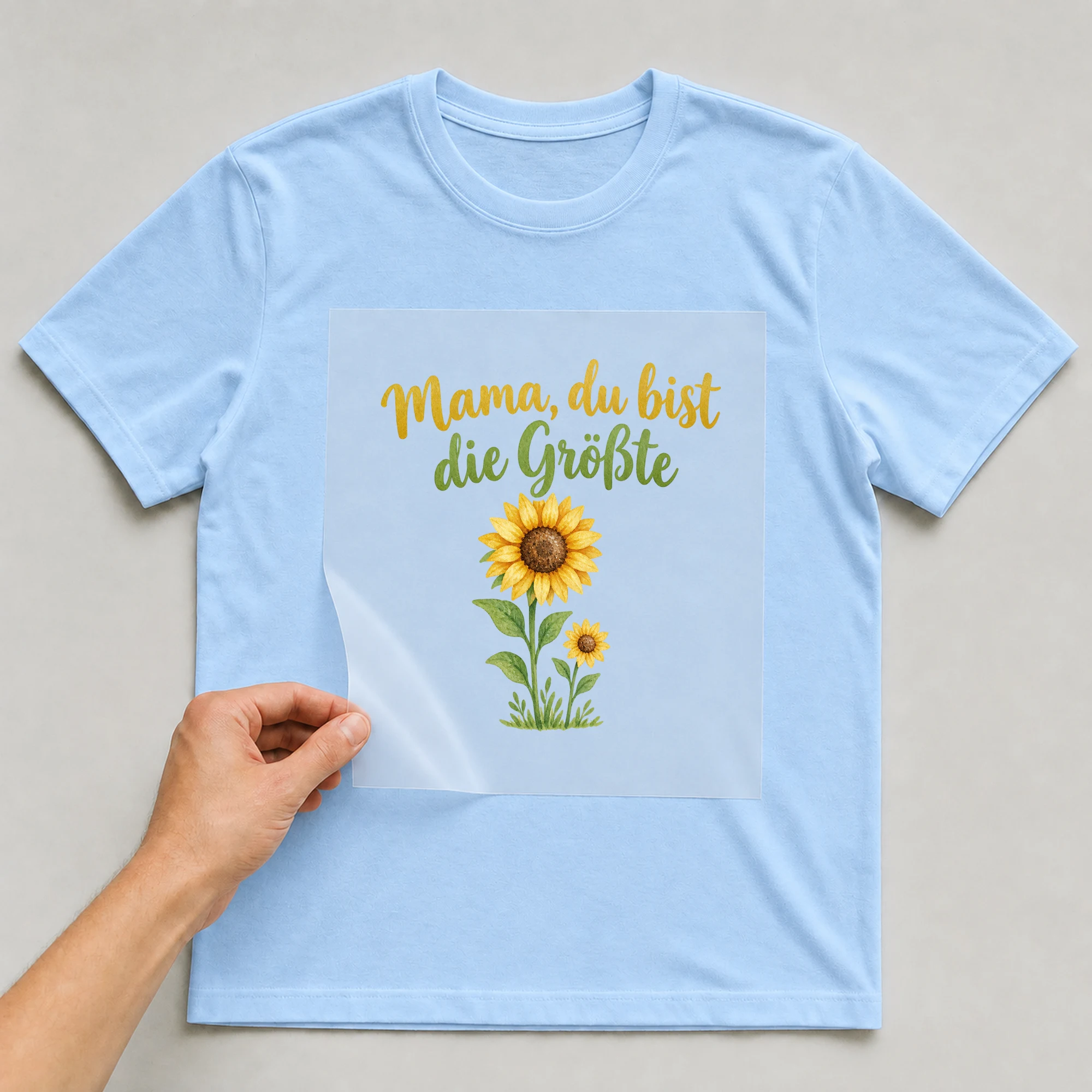 Bügelbild „Mama, du bist die Größte“ auf babyblauem T-Shirt, Folie wird aufgelegt