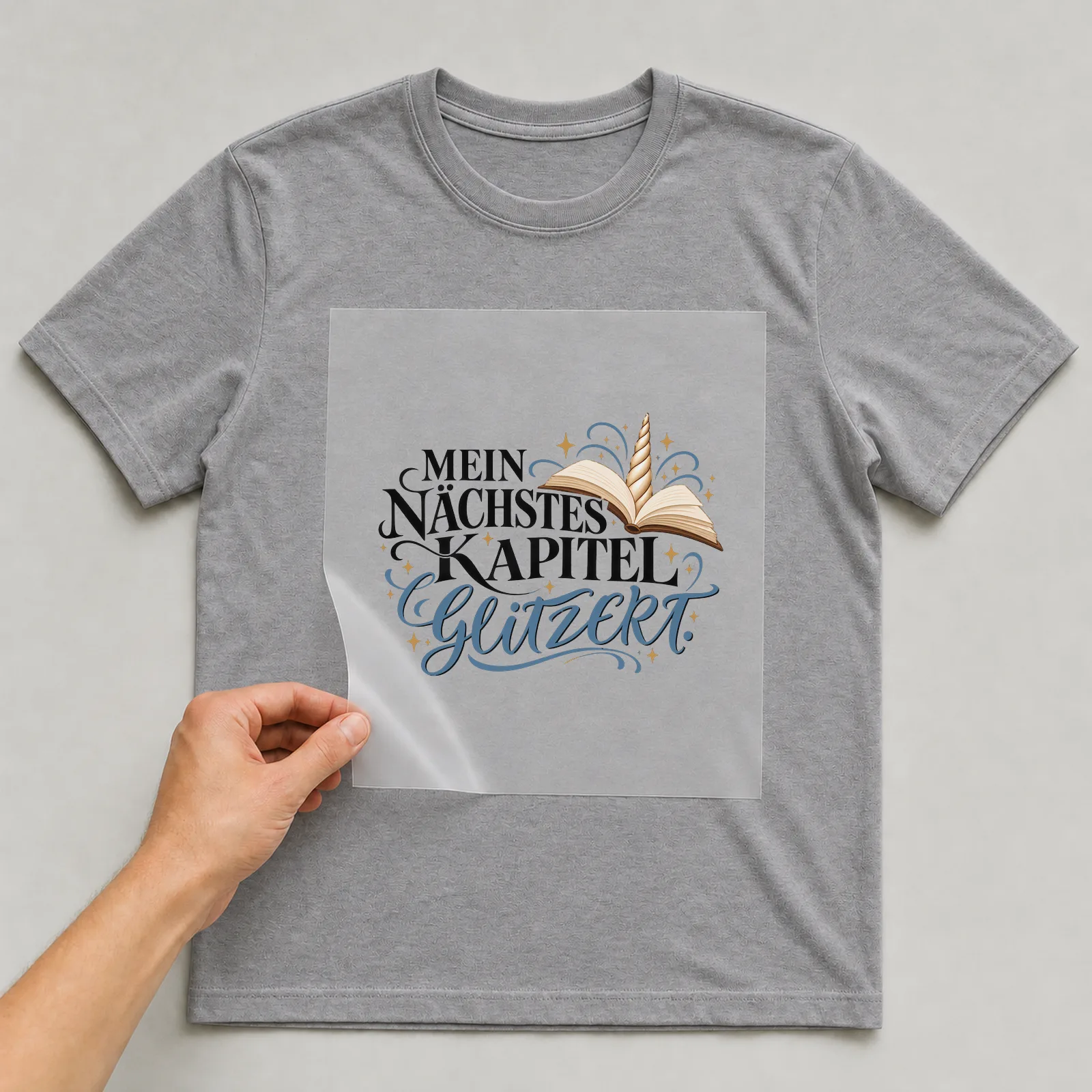 Bügelbild „Mein nächstes Kapitel glitzert“ auf grauem T-Shirt, Folie wird aufgelegt