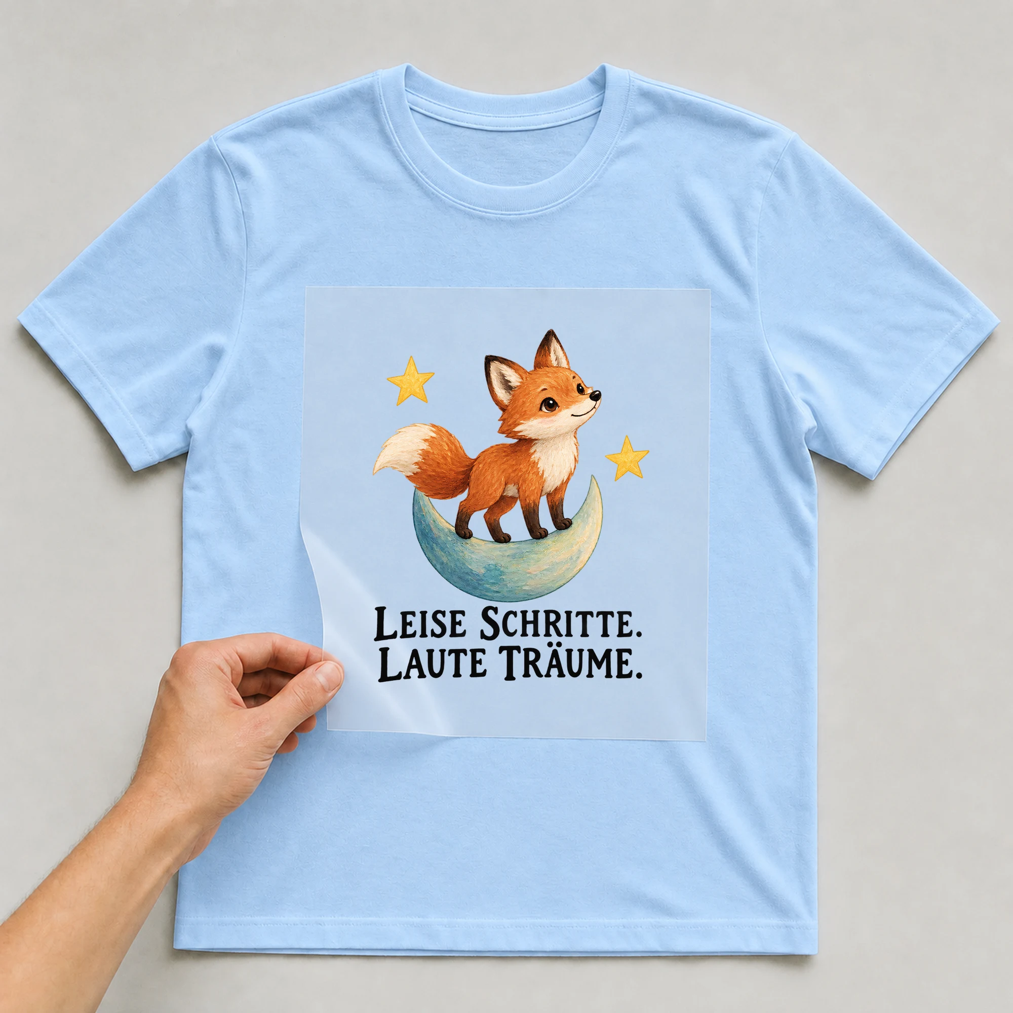 Bügelbild „LEISE SCHRITTE. LAUTE TRÄUME.“ auf babyblauem T-Shirt, Folie wird aufgelegt