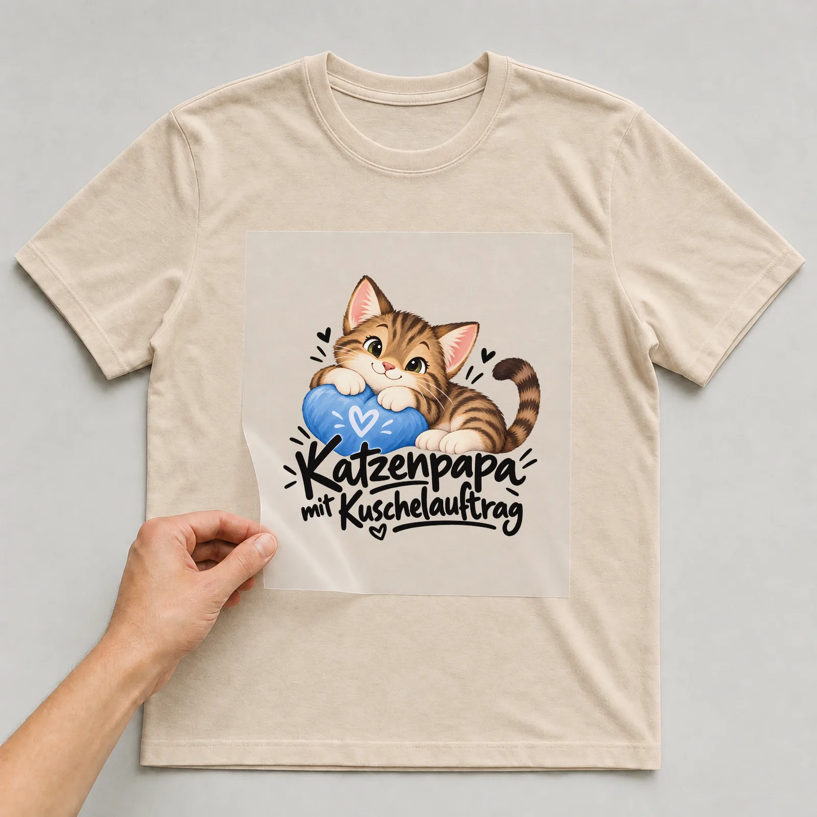Bügelbild „Katzenpapa mit Kuschelauftrag“ auf beigem T-Shirt, Folie wird aufgelegt