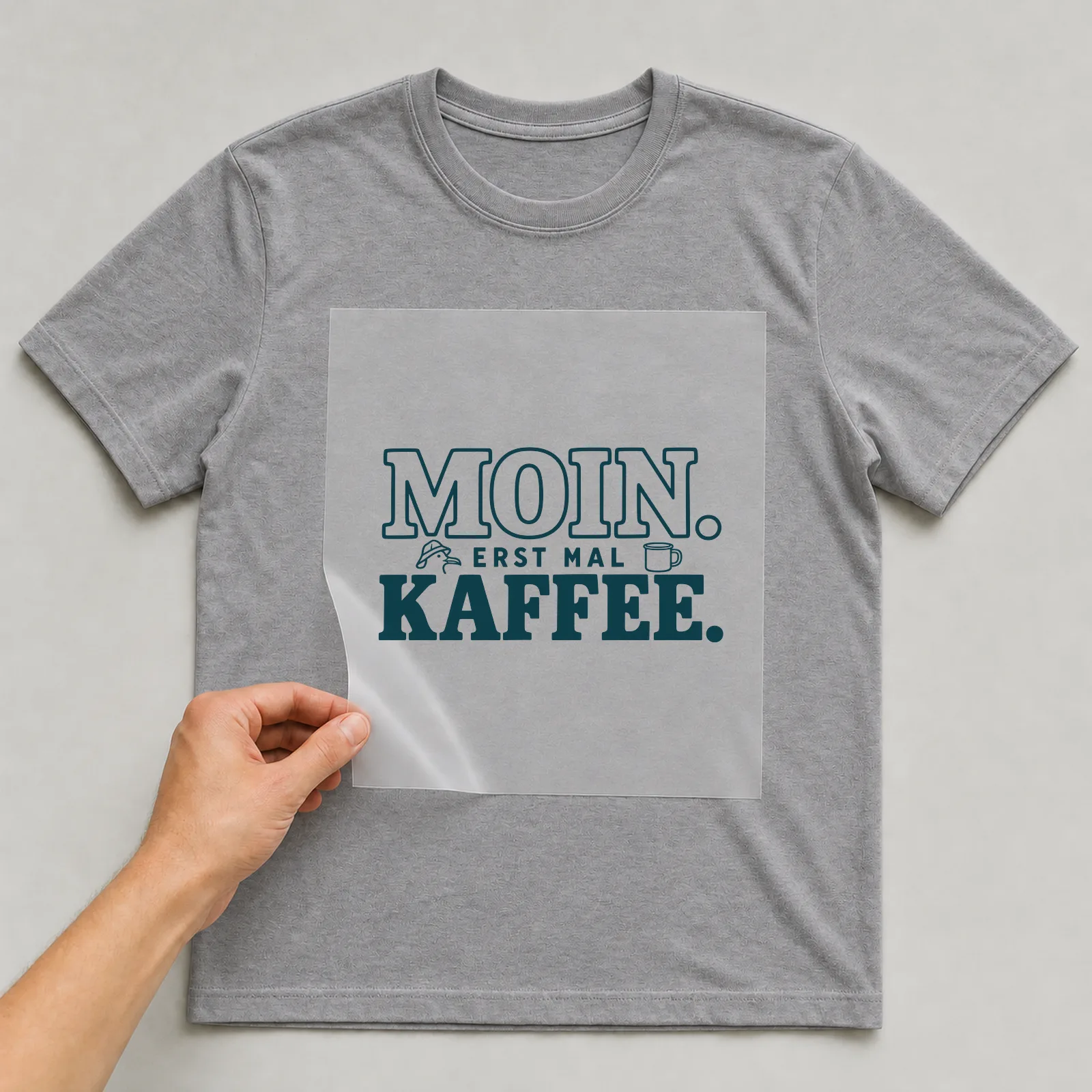 Bügelbild „Moin. Erst mal Kaffee.“ (Schrift Konturen) auf grauem T-Shirt, Folie wird aufgelegt