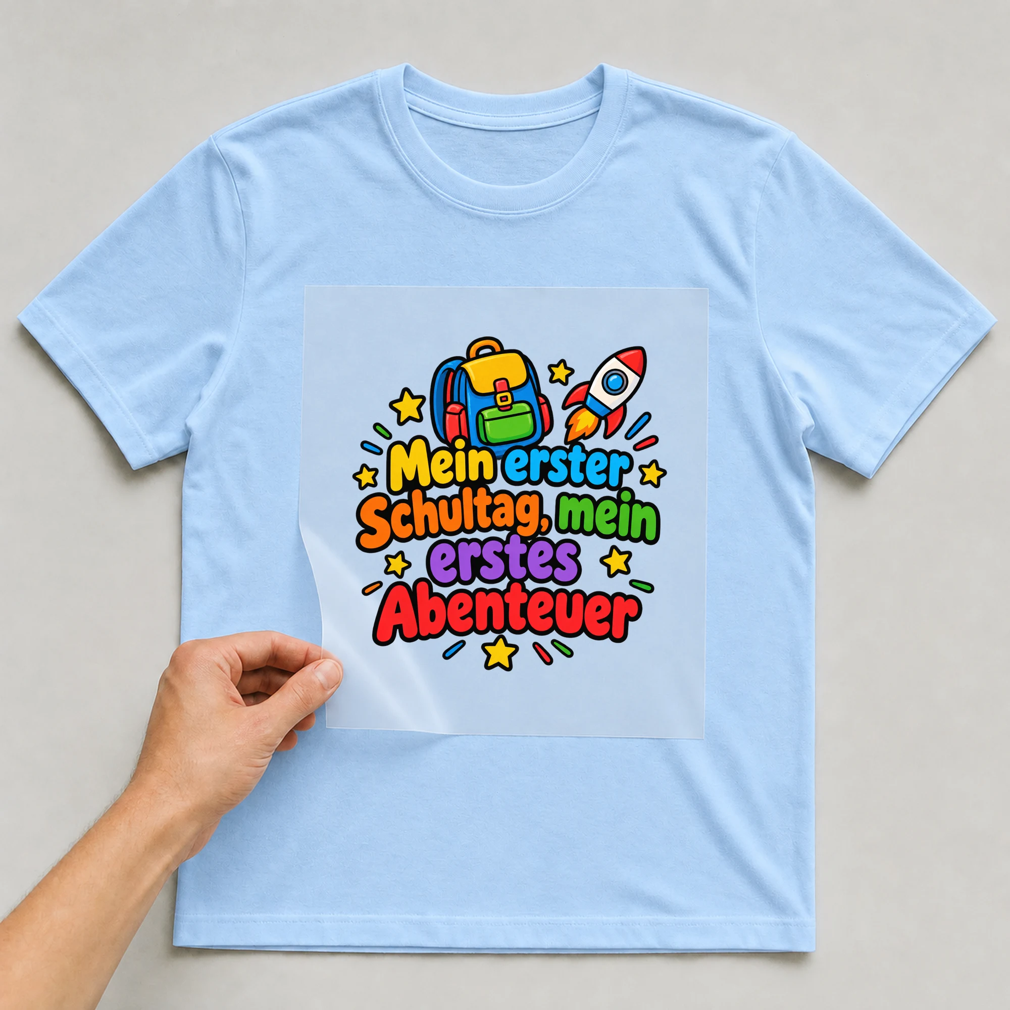 Bügelbild „Mein erster schultag mein erstes abenteuer“ auf babyblauem T-Shirt, Folie wird aufgelegt