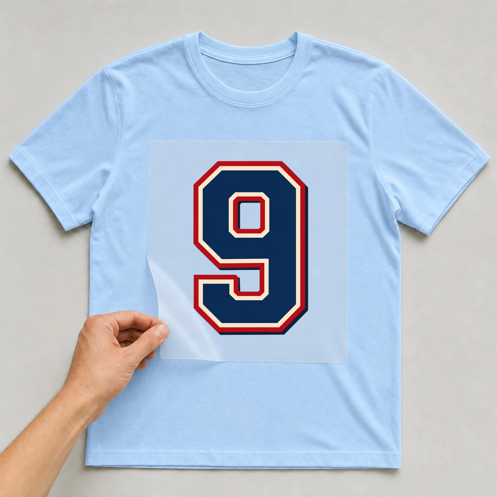 Bügelbild „Zahl 9 im Retro-College-Stil“ auf babyblauem T-Shirt, Folie wird aufgelegt
