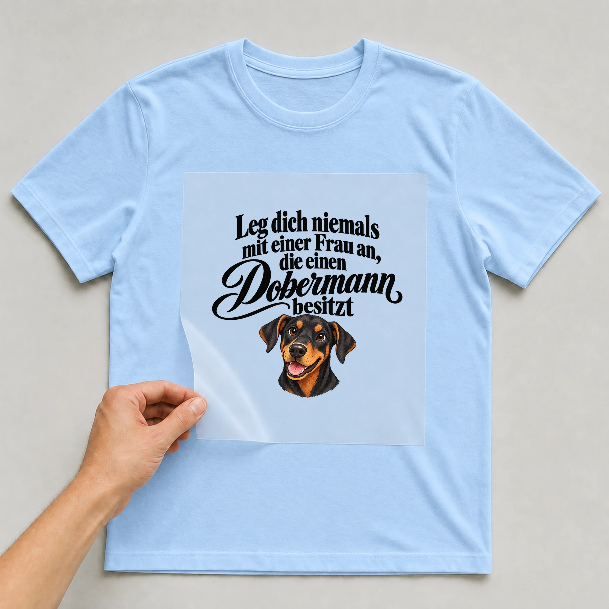 Bügelbild „Leg dich niemals mit einer Frau an, die einen Dobermann besitzt“ auf babyblauem T-Shirt, Folie wird aufgelegt