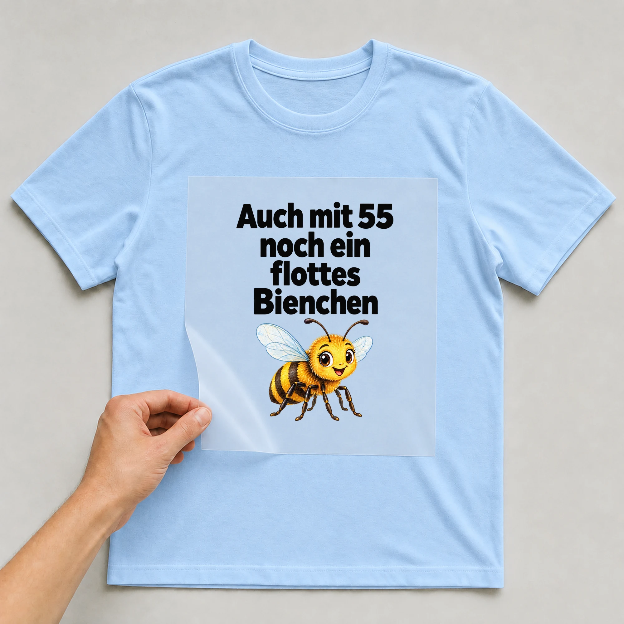 Bügelbild „Auch mit 55 noch ein flottes Bienchen“ auf babyblauem T-Shirt, Folie wird aufgelegt