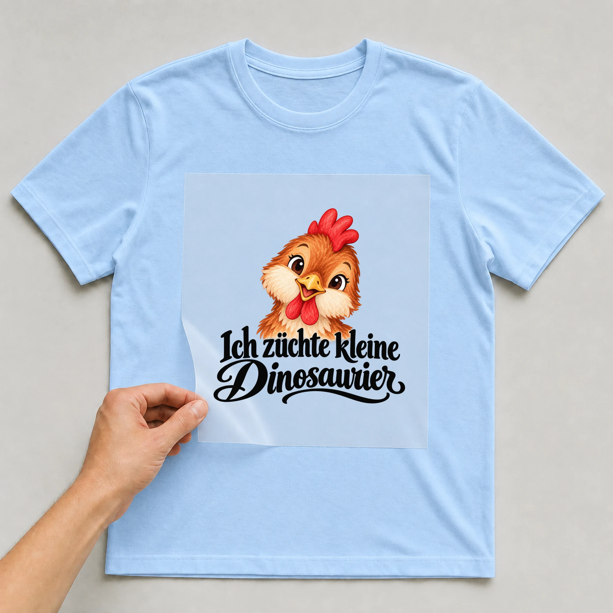 Bügelbild „Ich züchte kleine Dinosaurier“ auf babyblauem T-Shirt, Folie wird aufgelegt