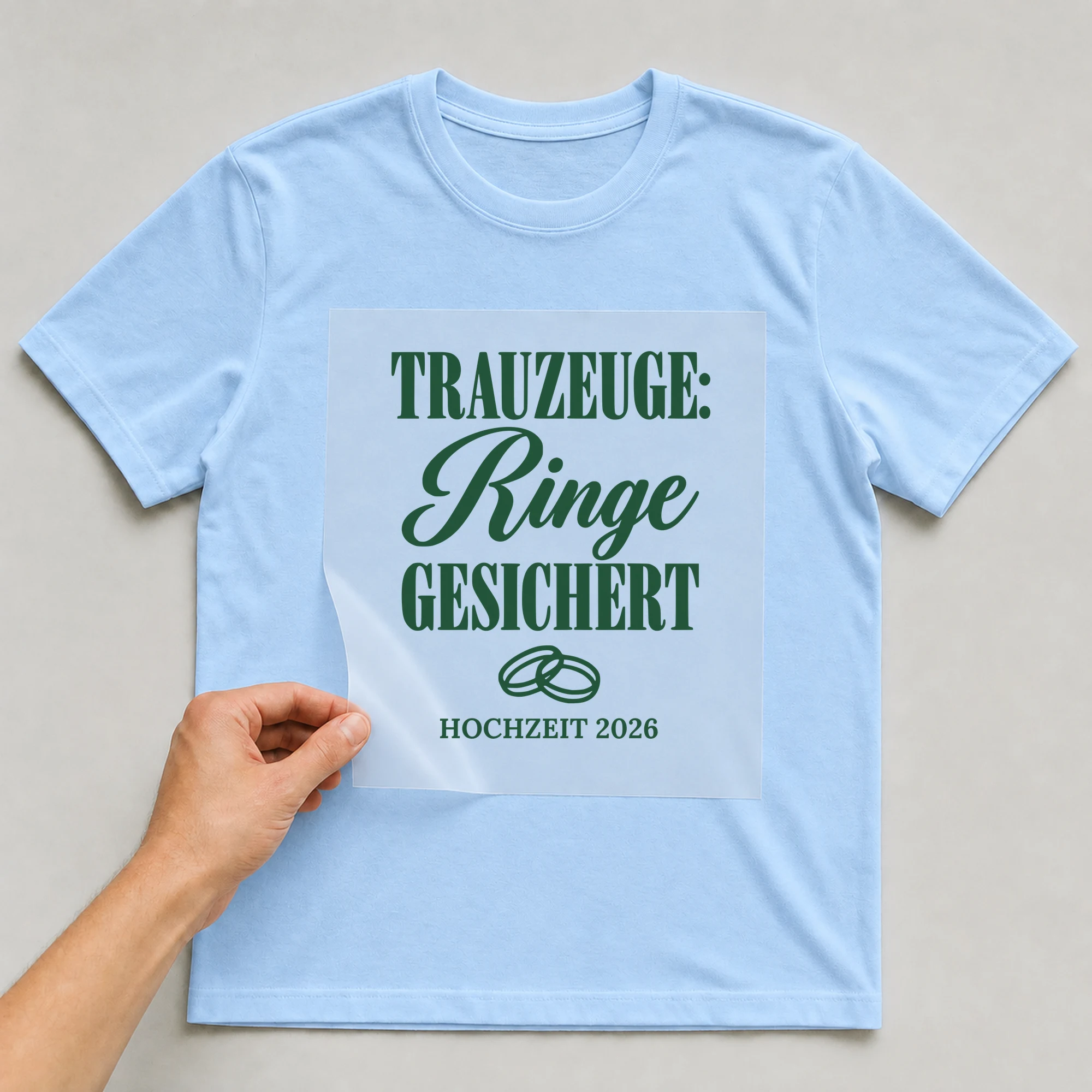 Bügelbild Trauzeuge: Ringe gesichert mit Jahr auf hellblauem T-Shirt, Folie wird aufgelegt