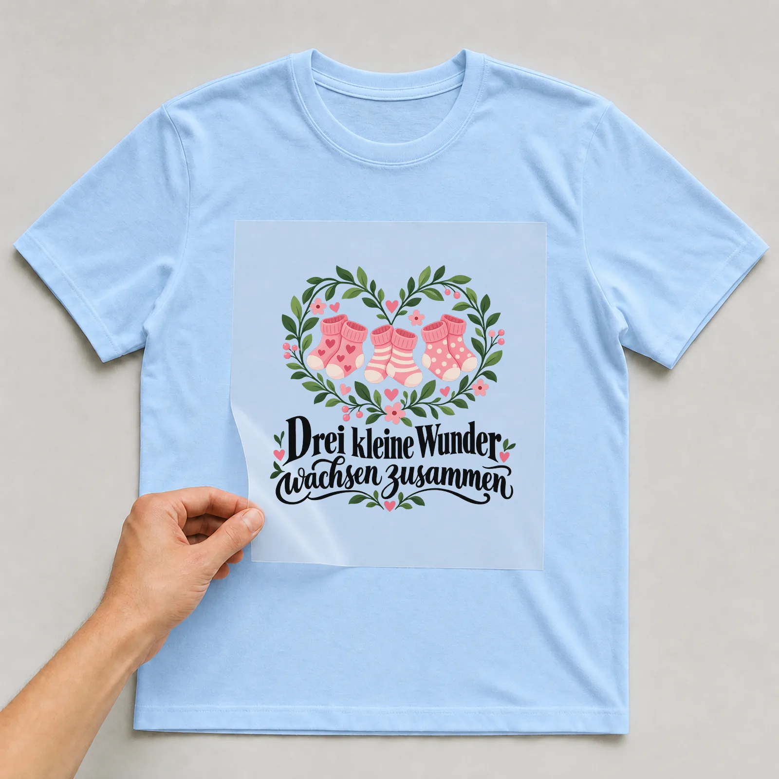 Bügelbild „Drei kleine Wunder wachsen zusammen“ auf hellblauem T-Shirt, Folie wird aufgelegt