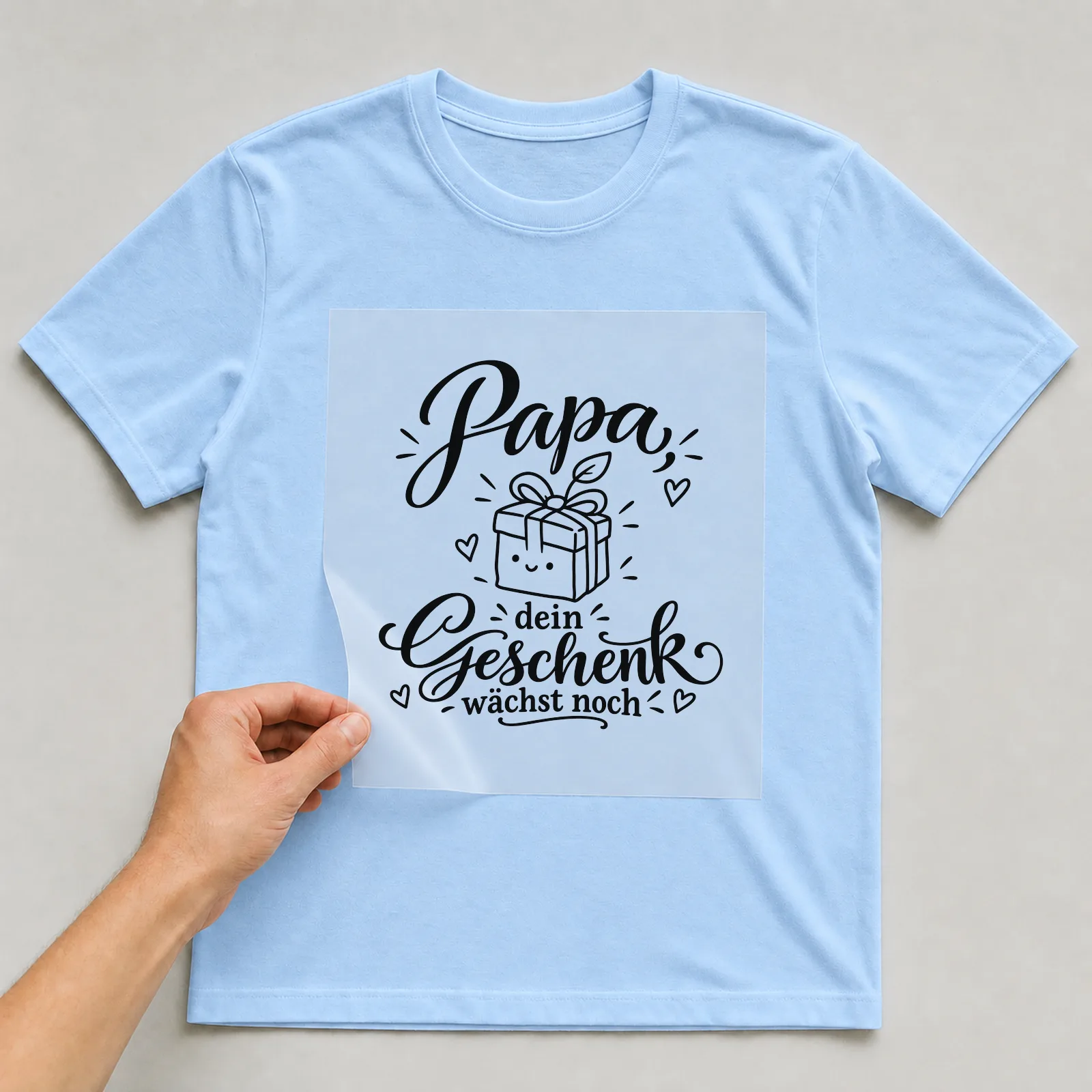 Bügelbild „Papa, dein Geschenk wächst noch“ auf hellblauem T-Shirt, Folie wird aufgelegt