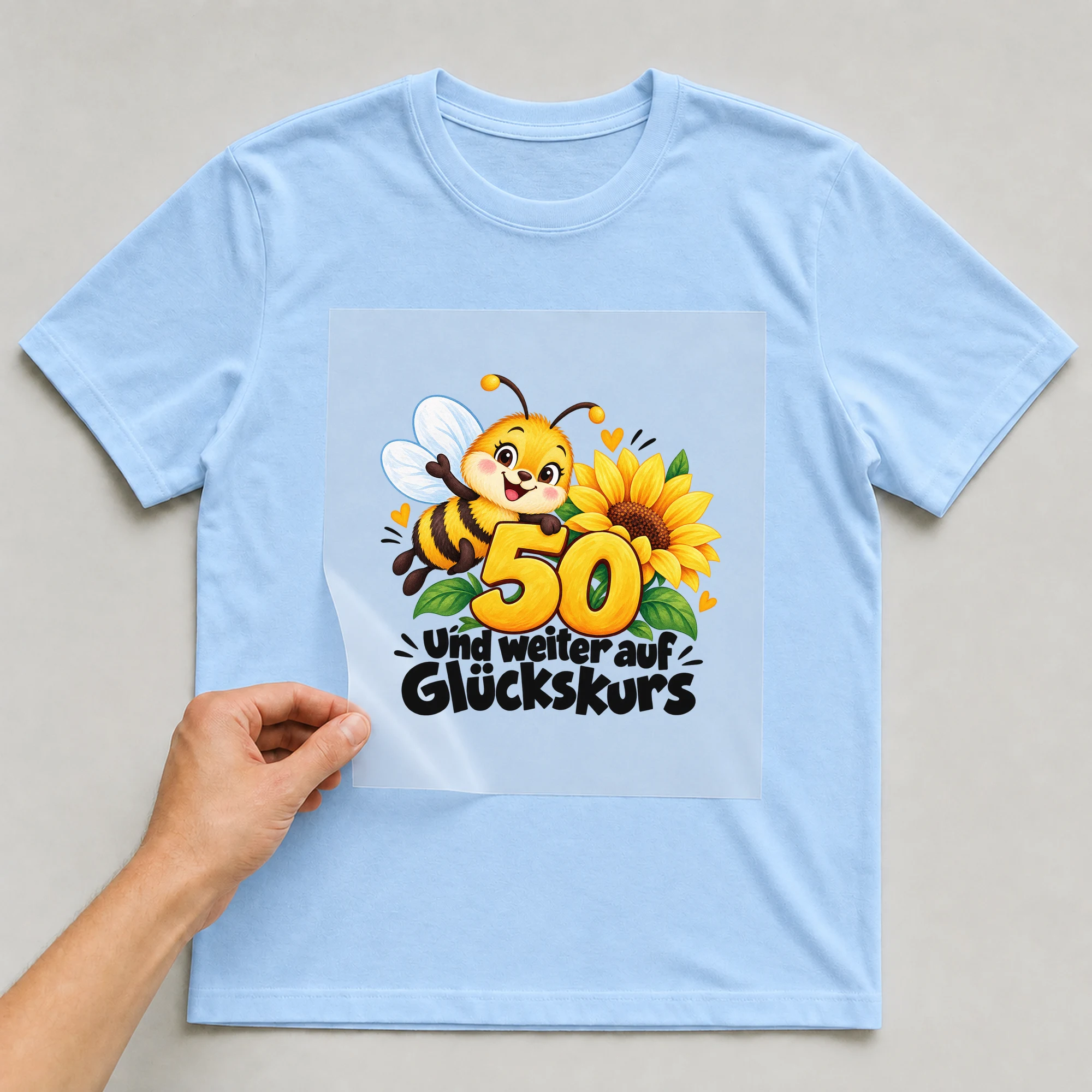 Bügelbild „50 und weiter auf Glückskurs“ auf babyblauem T-Shirt, Folie wird aufgelegt