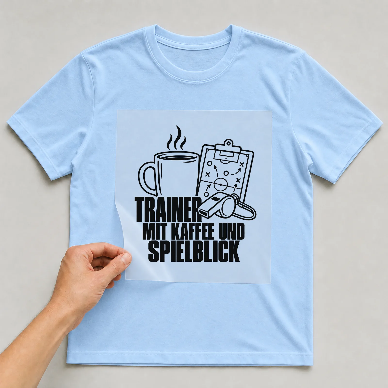 Bügelbild „Trainer mit Kaffee und Spielblick“ auf hellblauem T-Shirt, Folie wird aufgelegt