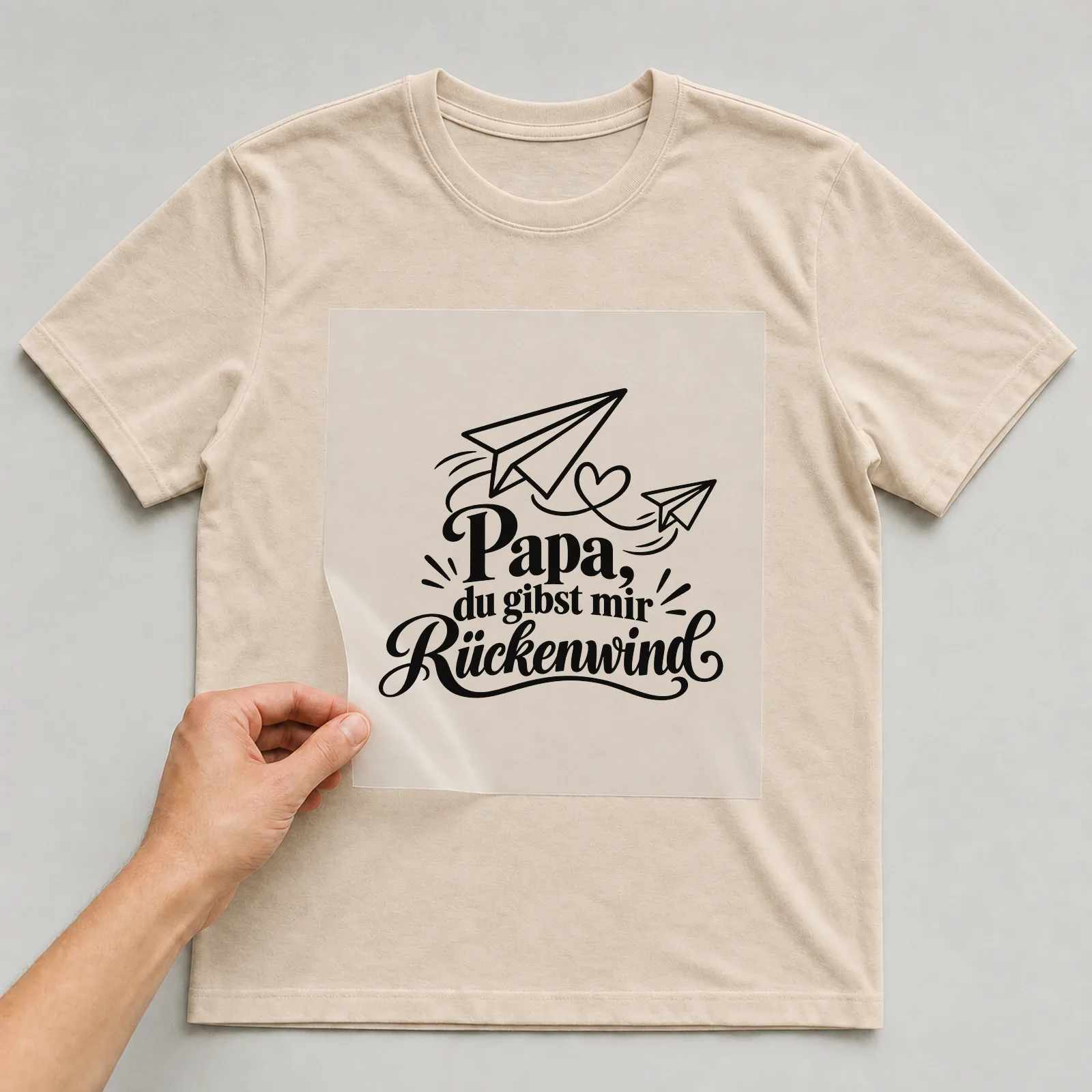Bügelbild „Papa, du gibst mir Rückenwind“ auf beigem T-Shirt, Folie wird aufgelegt