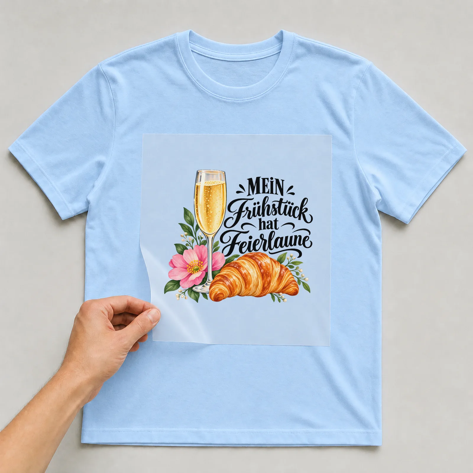 Bügelbild „MEIN Frühstück hat Feierlaune“ auf hellblauem T-Shirt, Folie wird aufgelegt