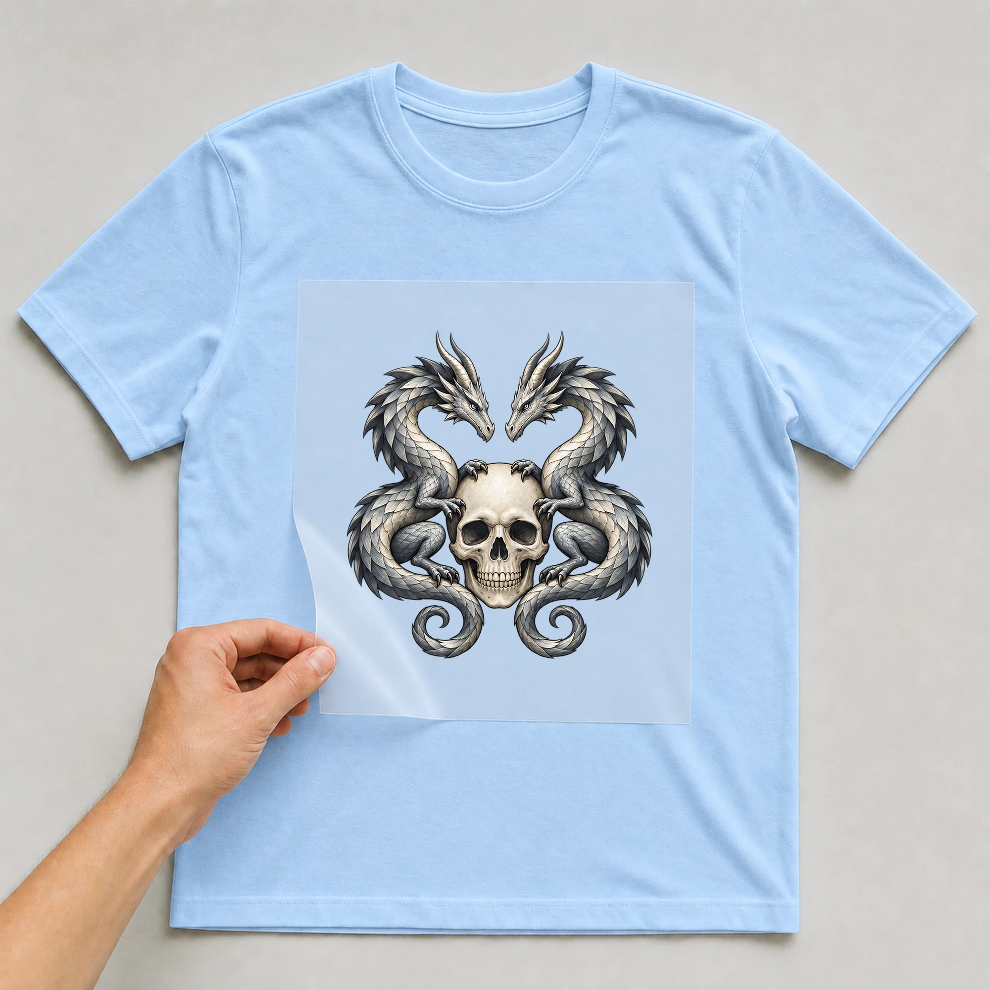Bügelbild „Totenkopf mit zwei Drachen“ auf babyblauem T-Shirt, Folie wird aufgelegt
