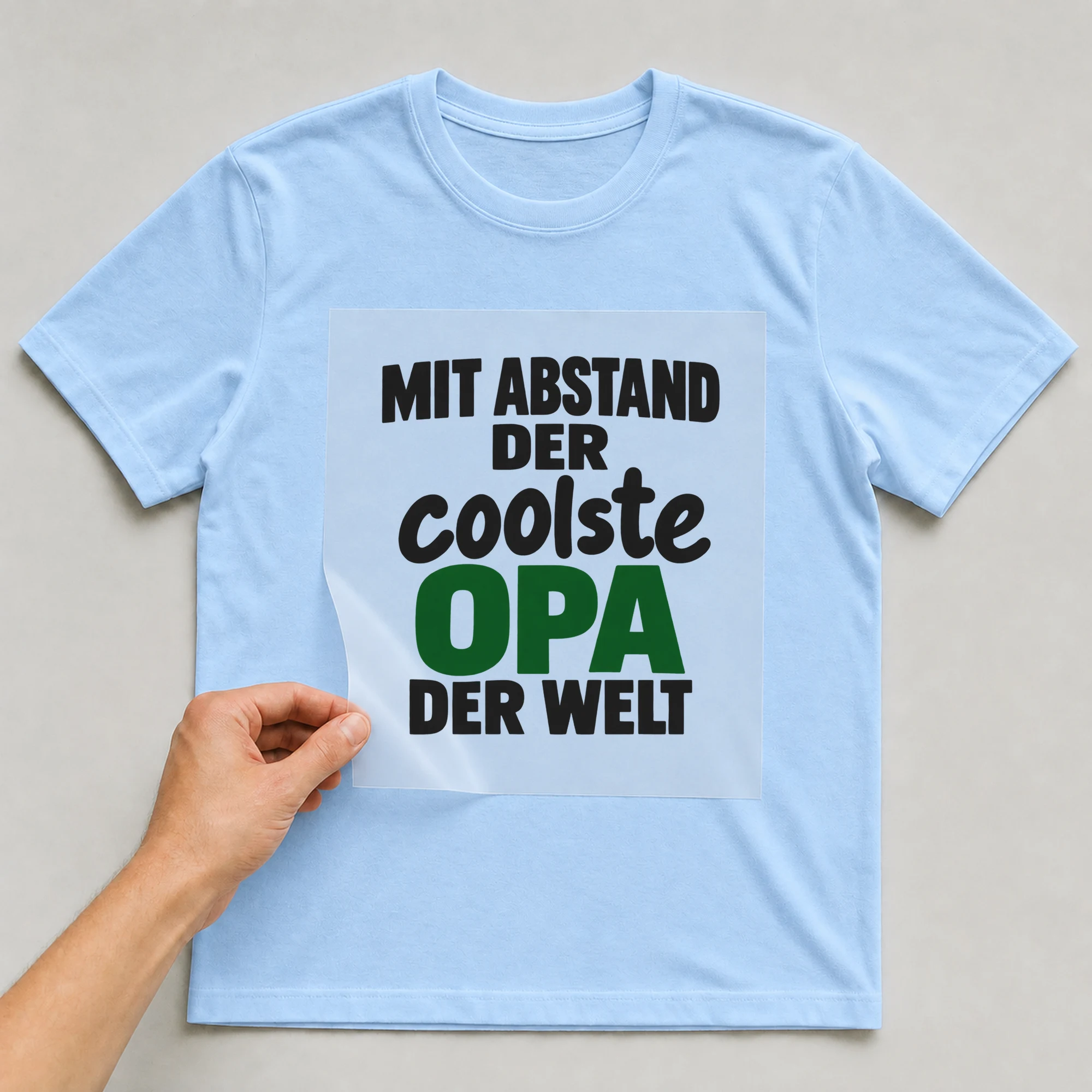 Bügelbild „Mit Abstand der coolste Opa der Welt“ auf babyblauem T-Shirt, Folie wird aufgelegt