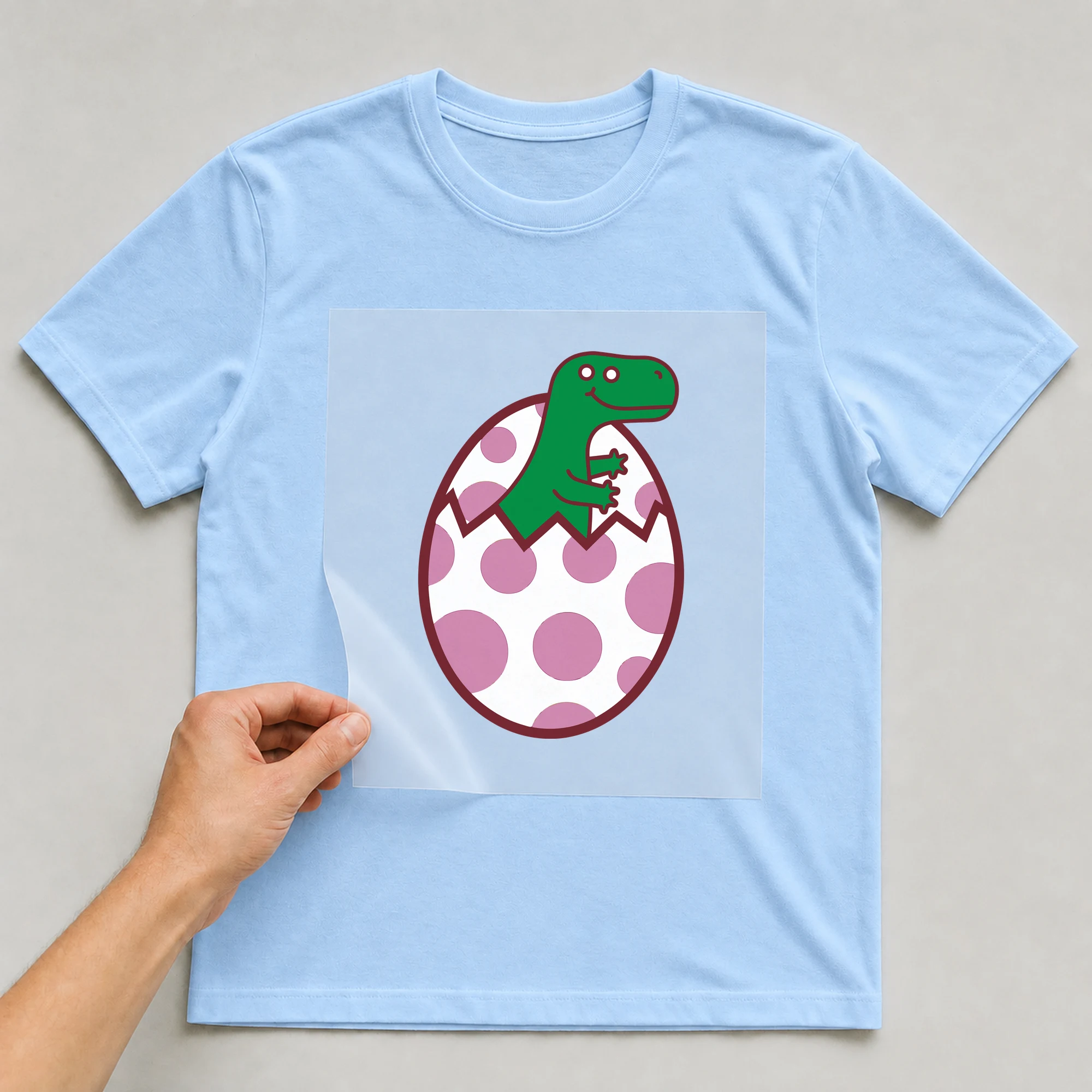 Bügelbild „Dino im Ei“ auf babyblauem T-Shirt, Folie wird aufgelegt