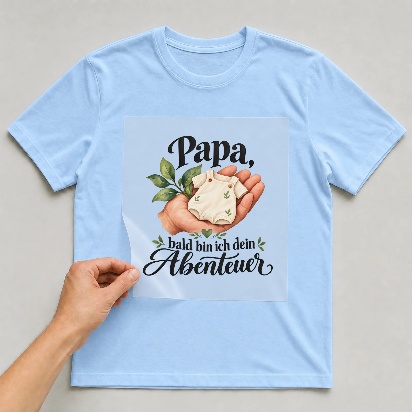 Bügelbild „Papa, bald bin ich dein Abenteuer“ auf hellblauem T-Shirt, Folie wird aufgelegt
