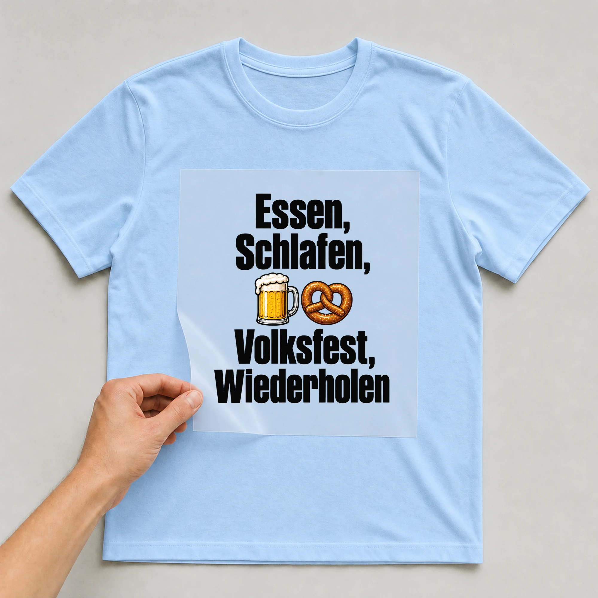 Bügelbild „Essen, Schlafen, Volksfest, Wiederholen“ auf babyblauem T-Shirt, Folie wird aufgelegt