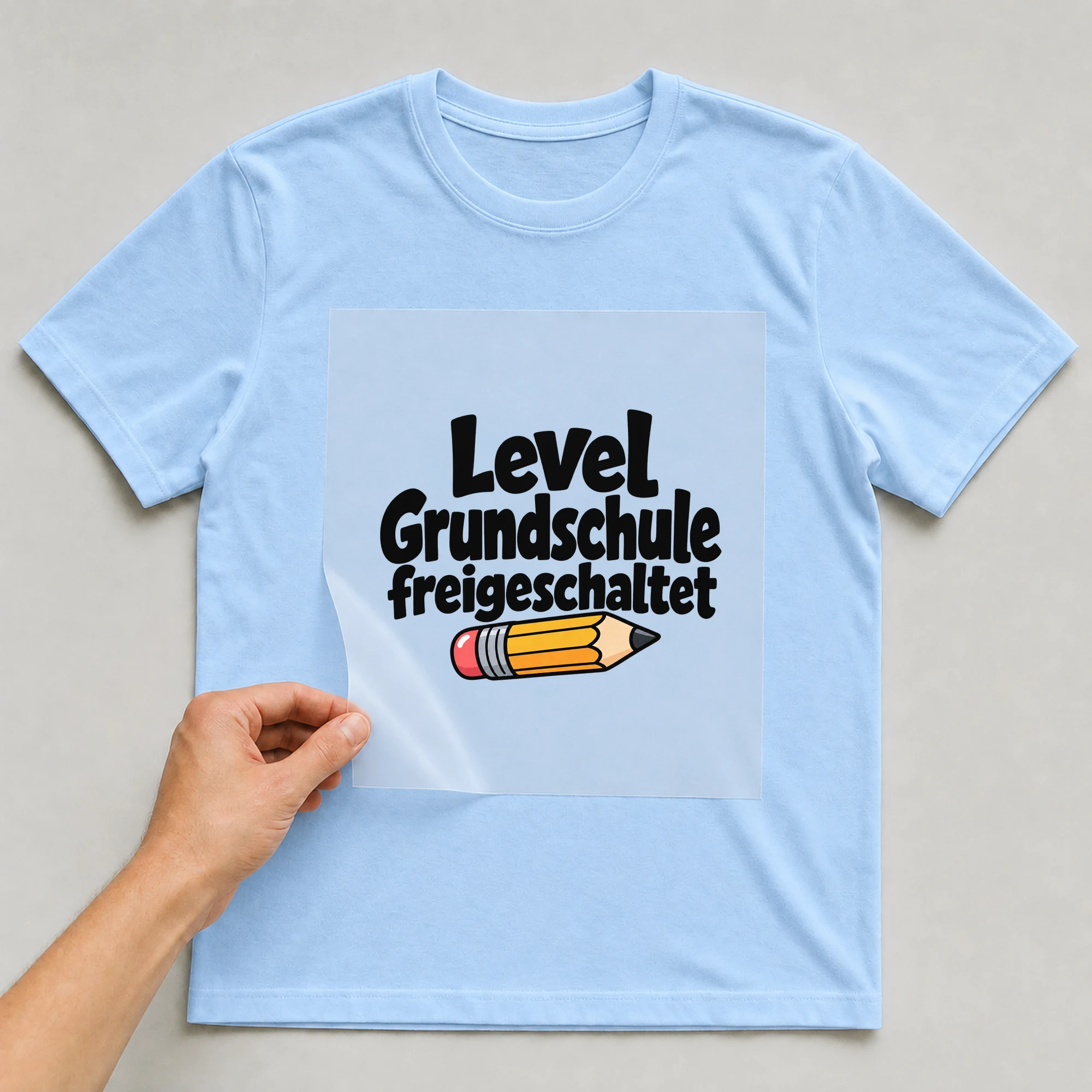 Bügelbild „Level Grundschule freigeschaltet“ auf babyblauem T-Shirt, Folie wird aufgelegt