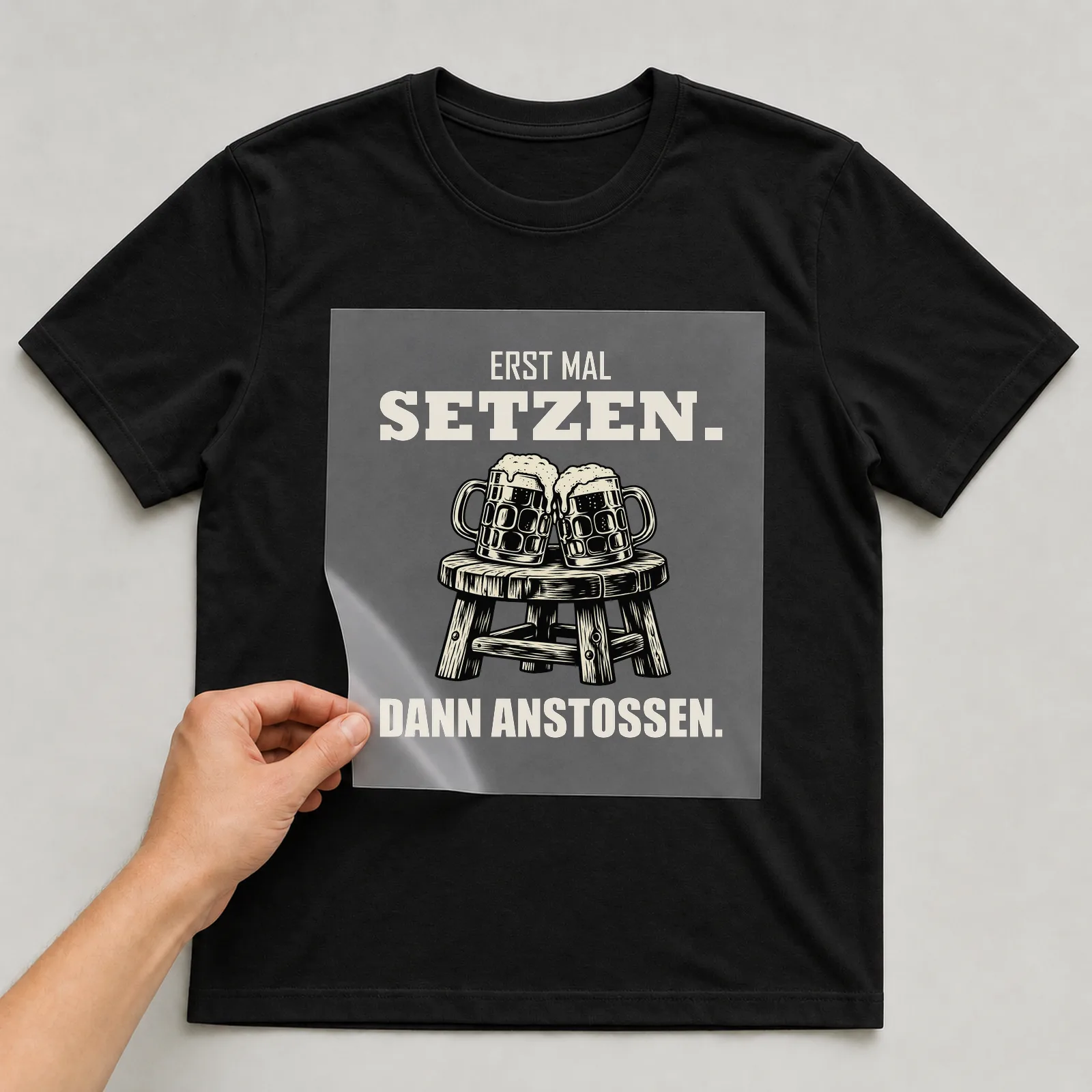Bügelbild „Erst mal setzen. Dann anstoßen.“ auf schwarzem T-Shirt, Folie wird aufgelegt