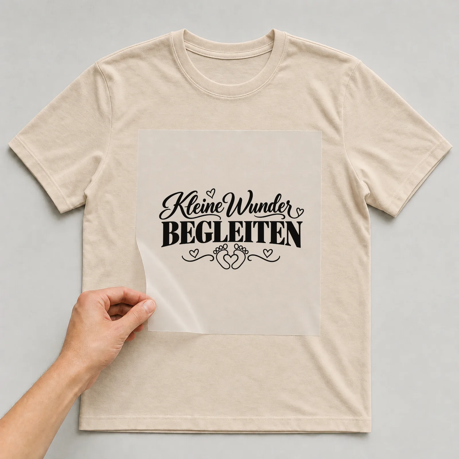 Bügelbild „Kleine Wunder begleiten“ auf beigem T-Shirt, Folie wird aufgelegt