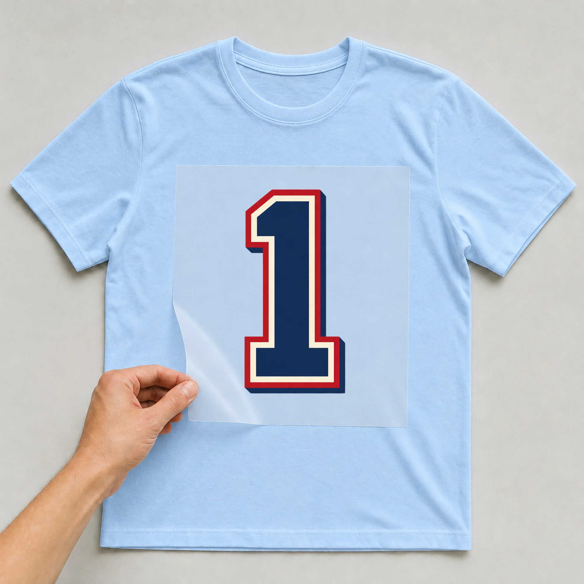 Bügelbild „Zahl 1 im Retro-College-Stil“ auf babyblauem T-Shirt, Folie wird aufgelegt