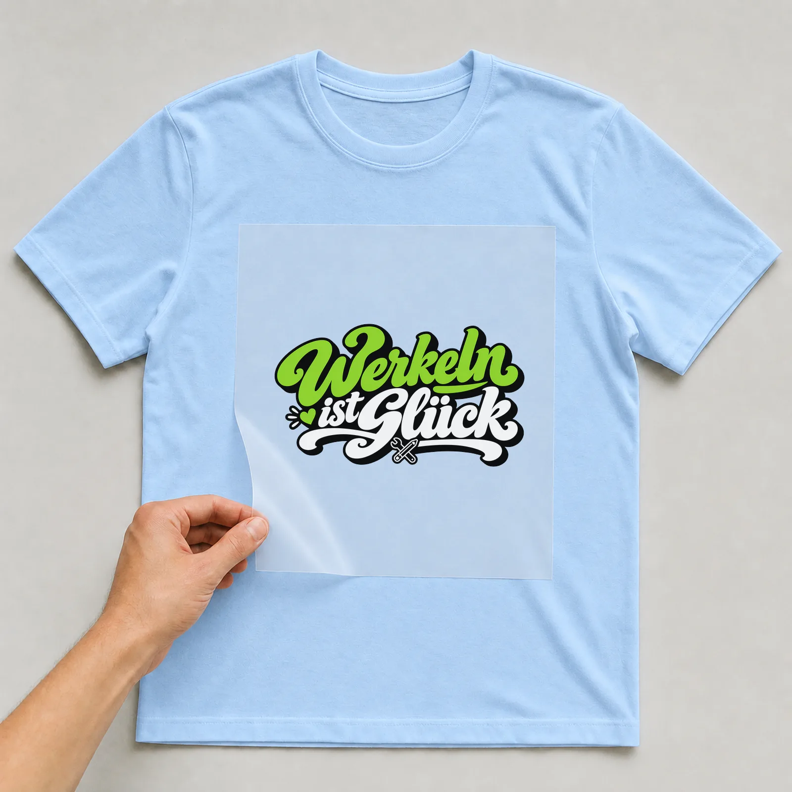 Bügelbild „Werkeln ist Glück“ auf hellblauem T-Shirt, Folie wird aufgelegt