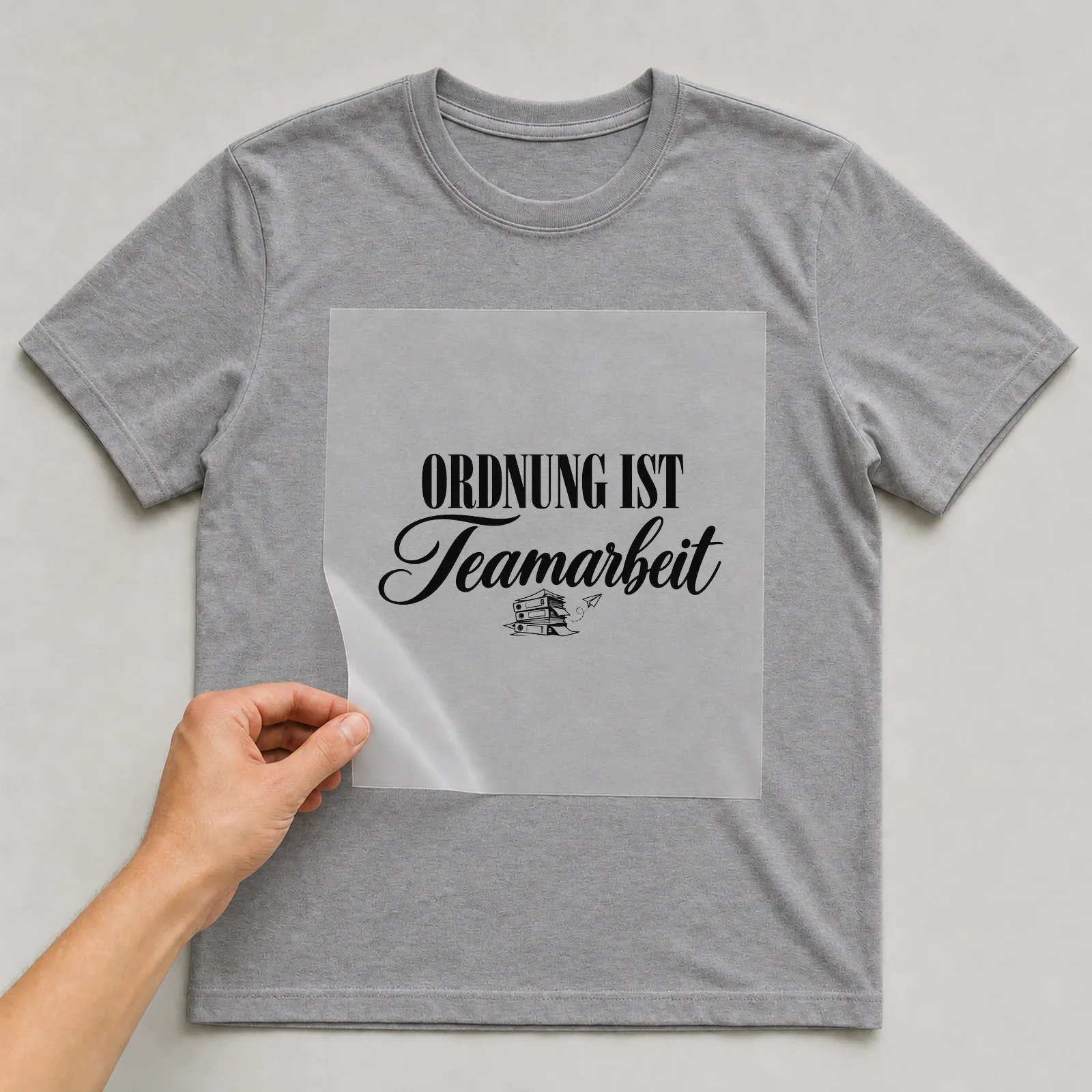 Bügelbild „Ordnung ist Teamarbeit“ auf grauem T-Shirt, Folie wird aufgelegt