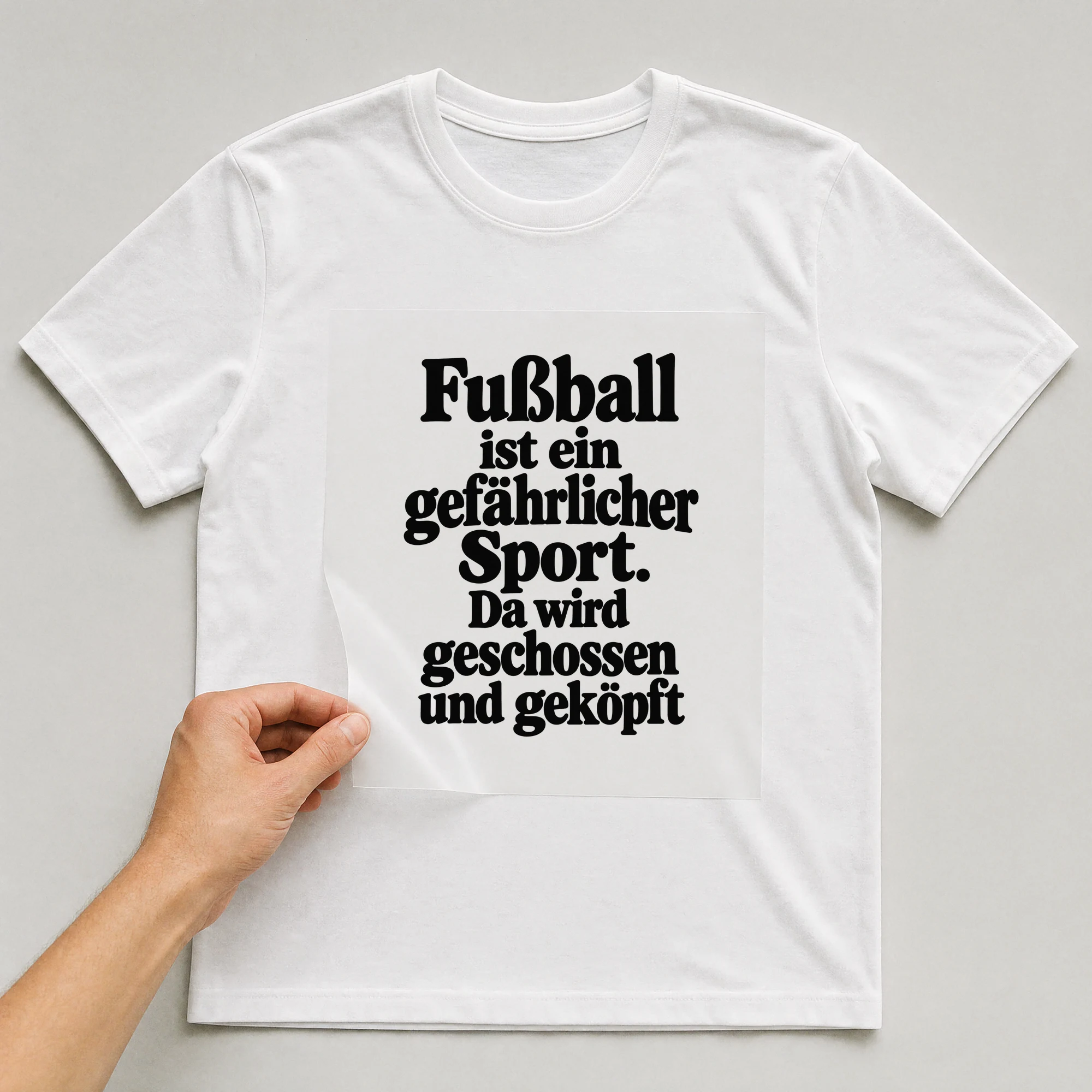 Bügelbild „Fußball ist ein gefährlicher Sport. Da wird geschossen und geköpft“ auf weißem T-Shirt, Folie wird aufgelegt
