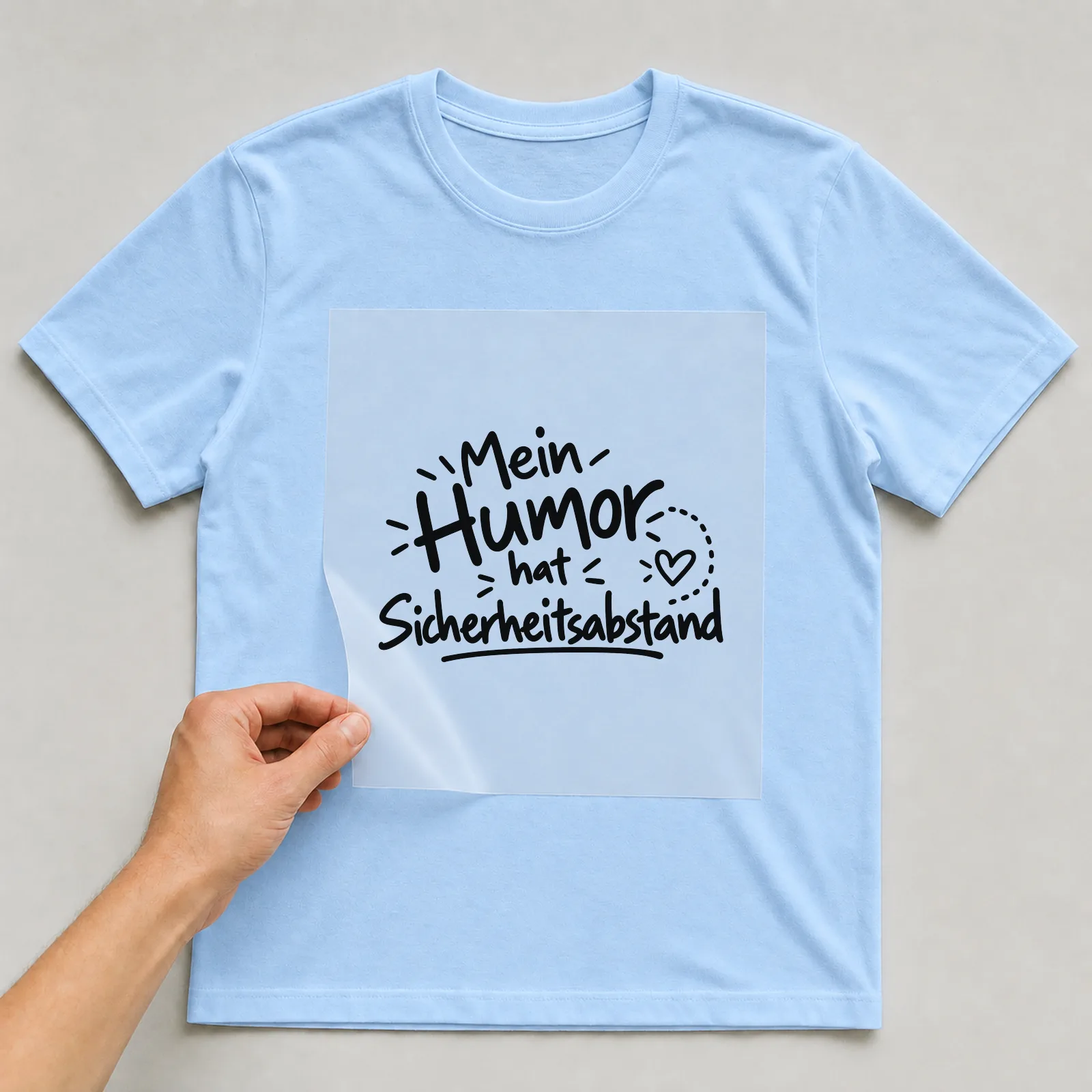 Bügelbild „Mein Humor hat Sicherheitsabstand“ auf hellblauem T-Shirt, Folie wird aufgelegt