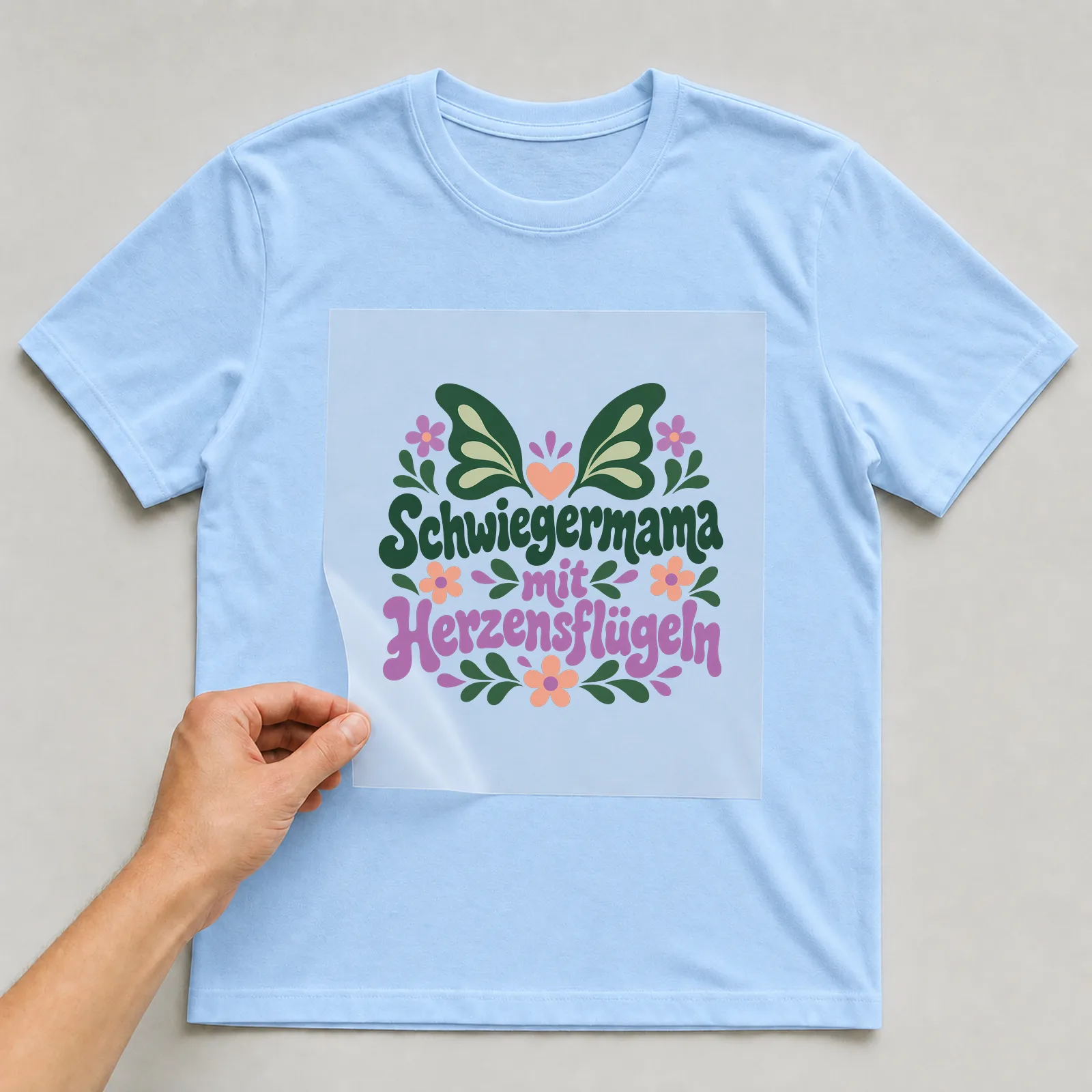 Bügelbild „Schwiegermama mit Herzensflügeln“ auf hellblauem T-Shirt, Folie wird aufgelegt