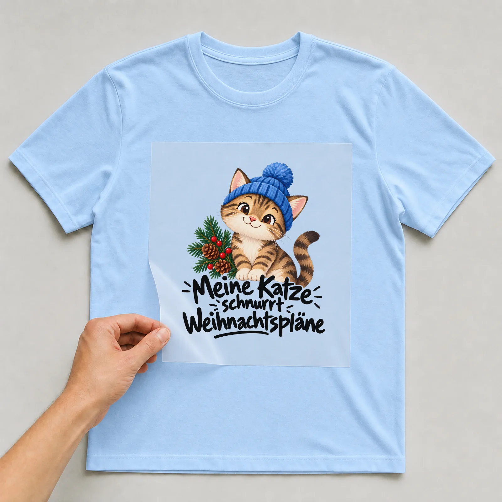 Bügelbild „Meine Katze schnurrt Weihnachtspläne“ auf hellblauem T-Shirt, Folie wird aufgelegt