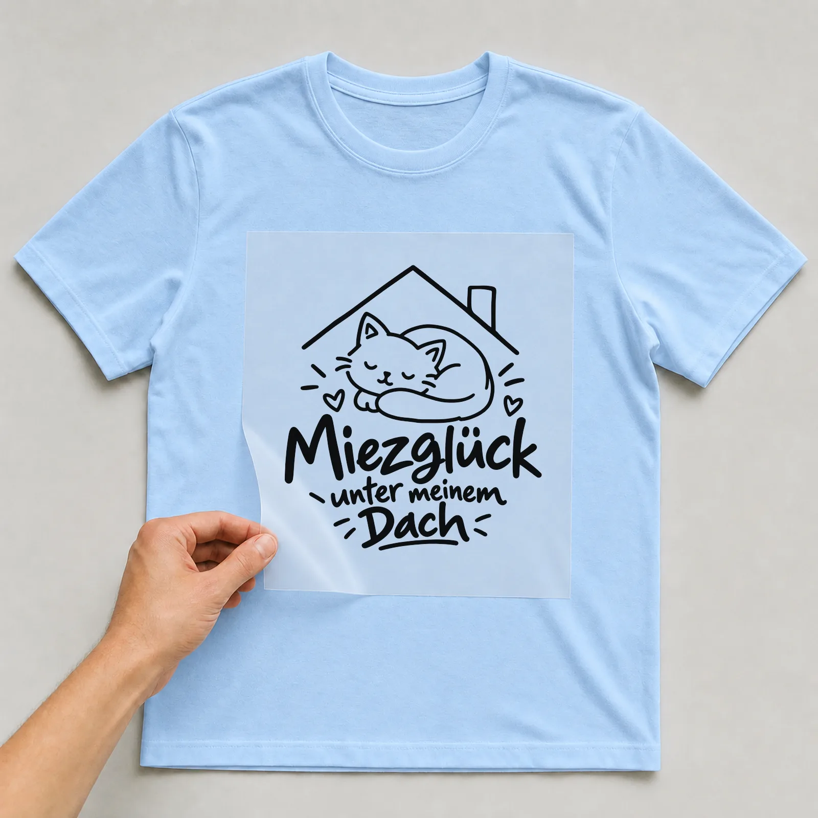 Bügelbild „Miezglück unter meinem Dach“ auf hellblauem T-Shirt, Folie wird aufgelegt