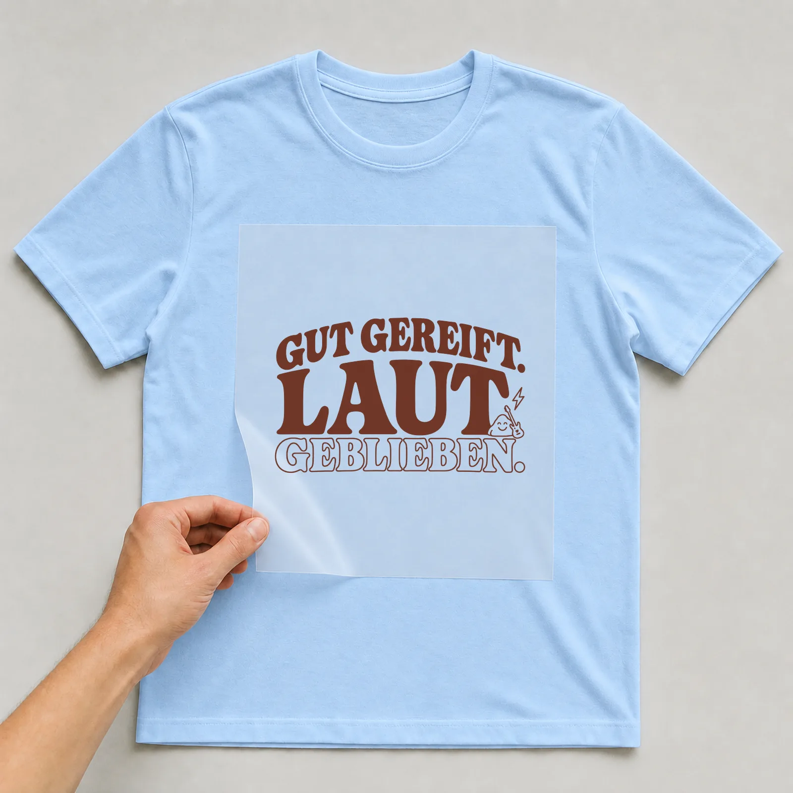 Bügelbild „Gut gereift. Laut geblieben.“ (Schrift Konturen) auf hellblauem T-Shirt, Folie wird aufgelegt