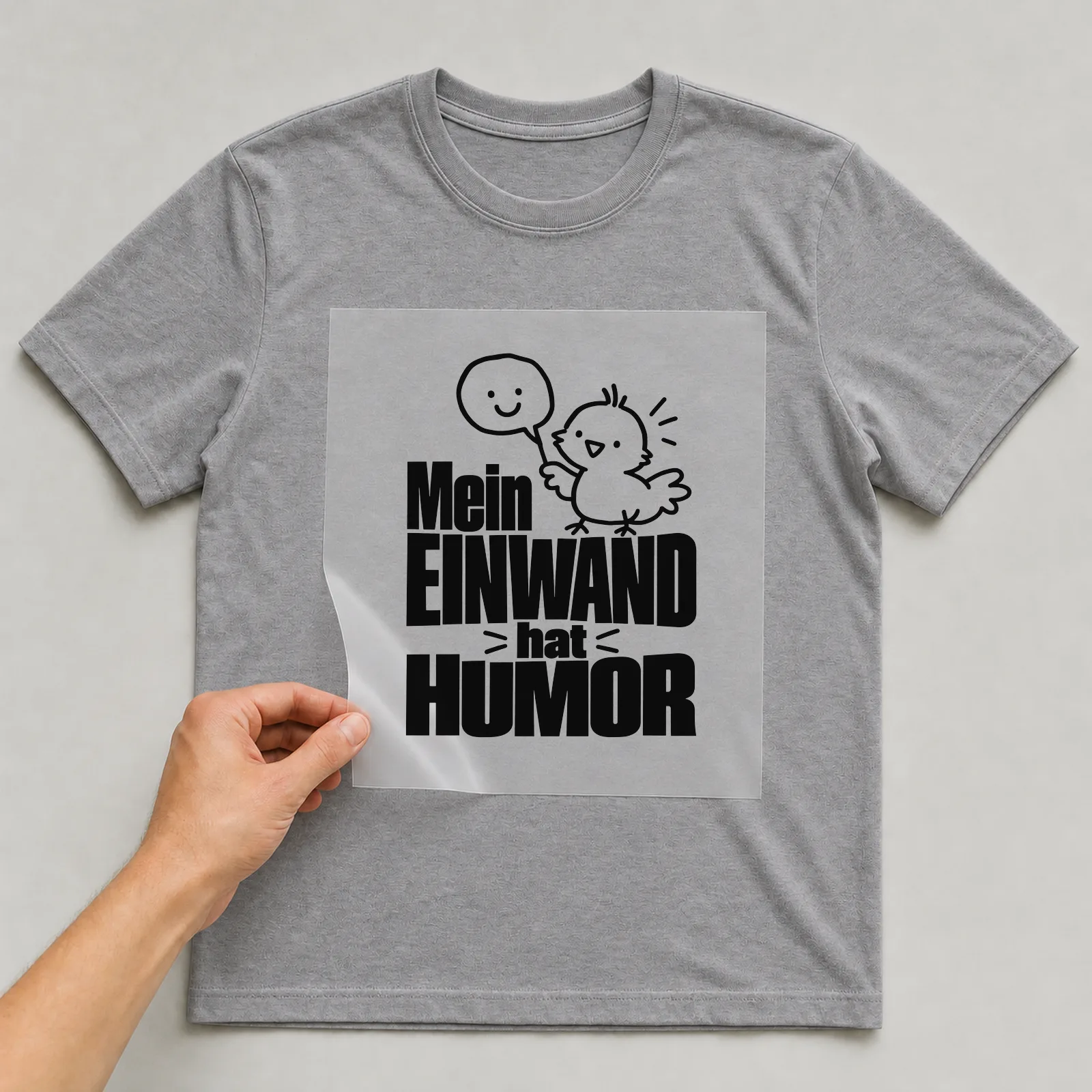 Bügelbild „Mein Einwand hat Humor“ auf grauem T-Shirt, Folie wird aufgelegt