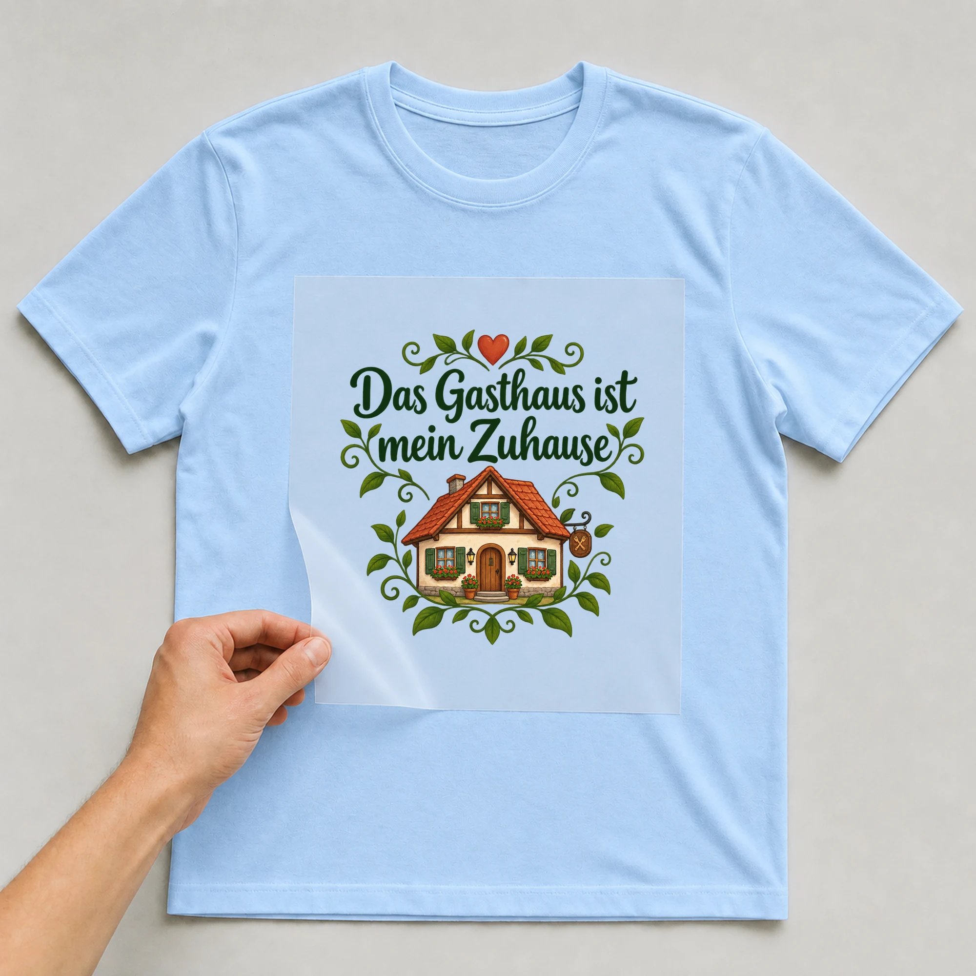 Bügelbild „Das Gasthaus ist mein Zuhause“ auf babyblauem T-Shirt, Folie wird aufgelegt