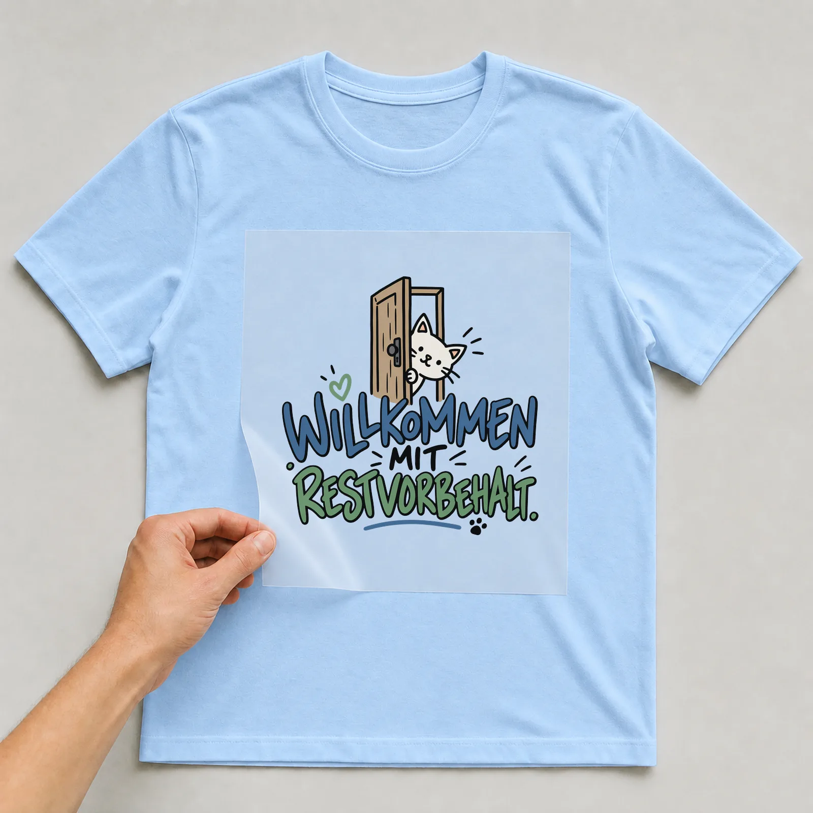 Bügelbild „Willkommen mit Restvorbehalt“ auf hellblauem T-Shirt, Folie wird aufgelegt