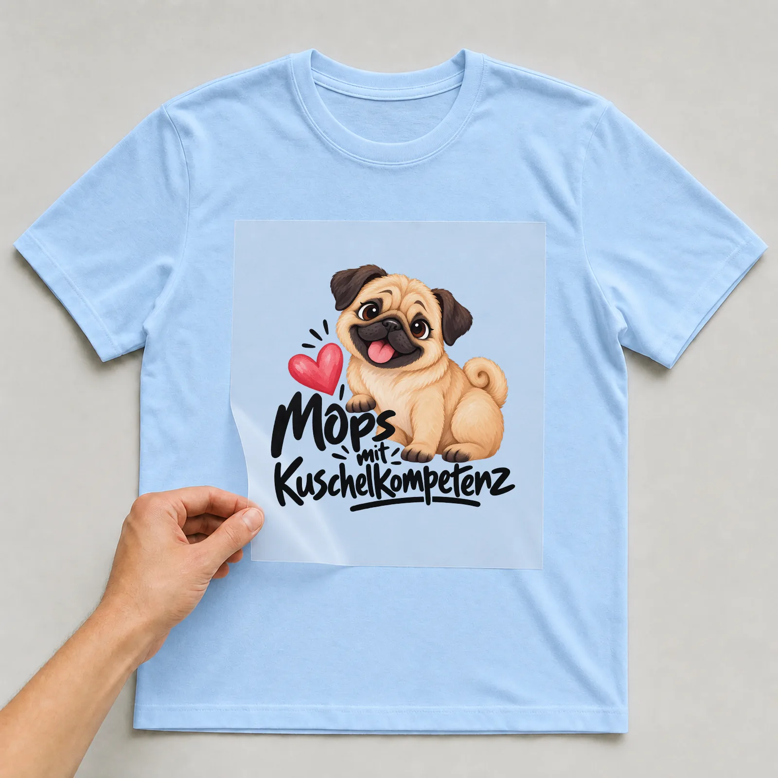 Bügelbild „Mops mit Kuschelkompetenz“ auf hellblauem T-Shirt, Folie wird aufgelegt