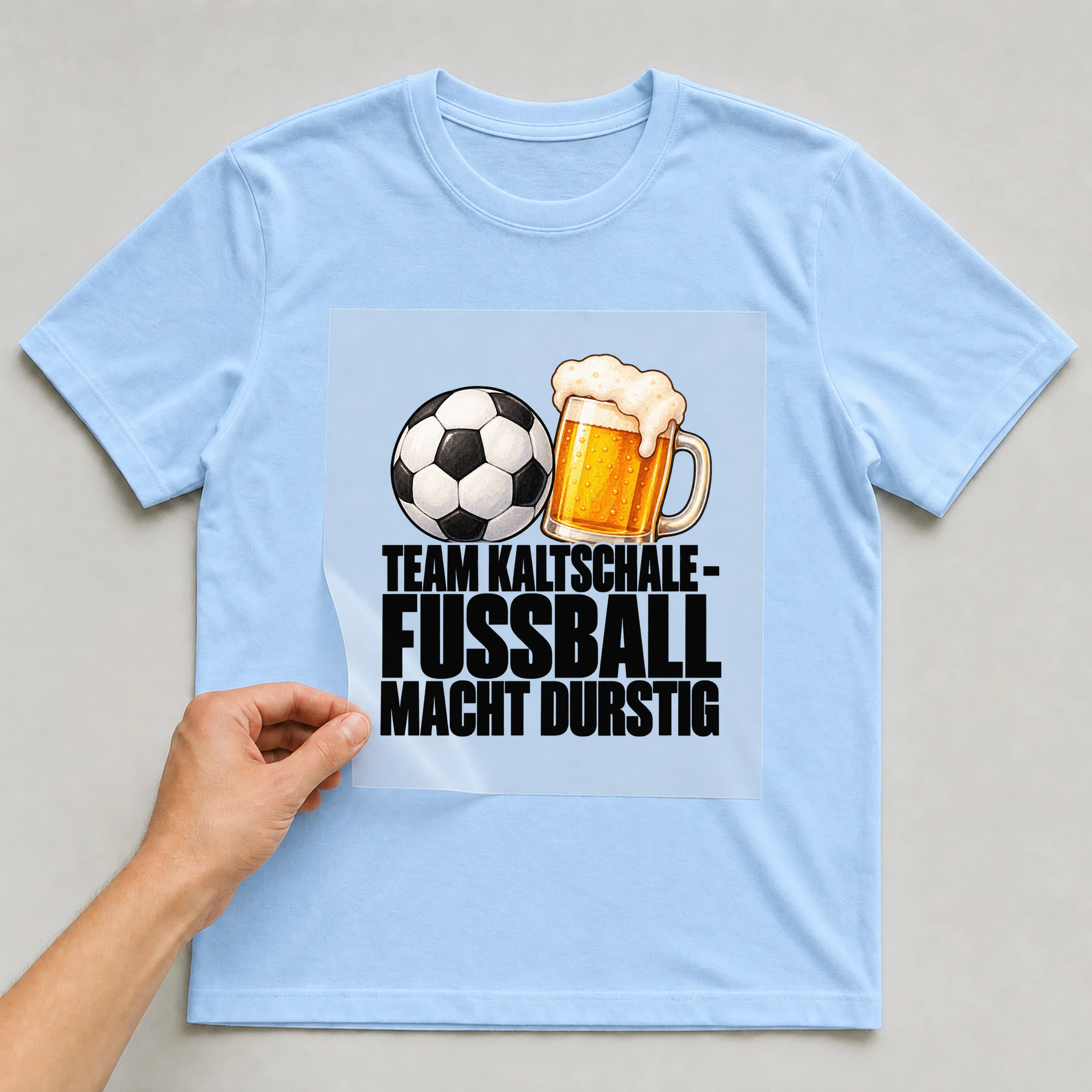Bügelbild „Team Kaltschale - Fussball Macht Durstig“ auf babyblauem T-Shirt, Folie wird aufgelegt