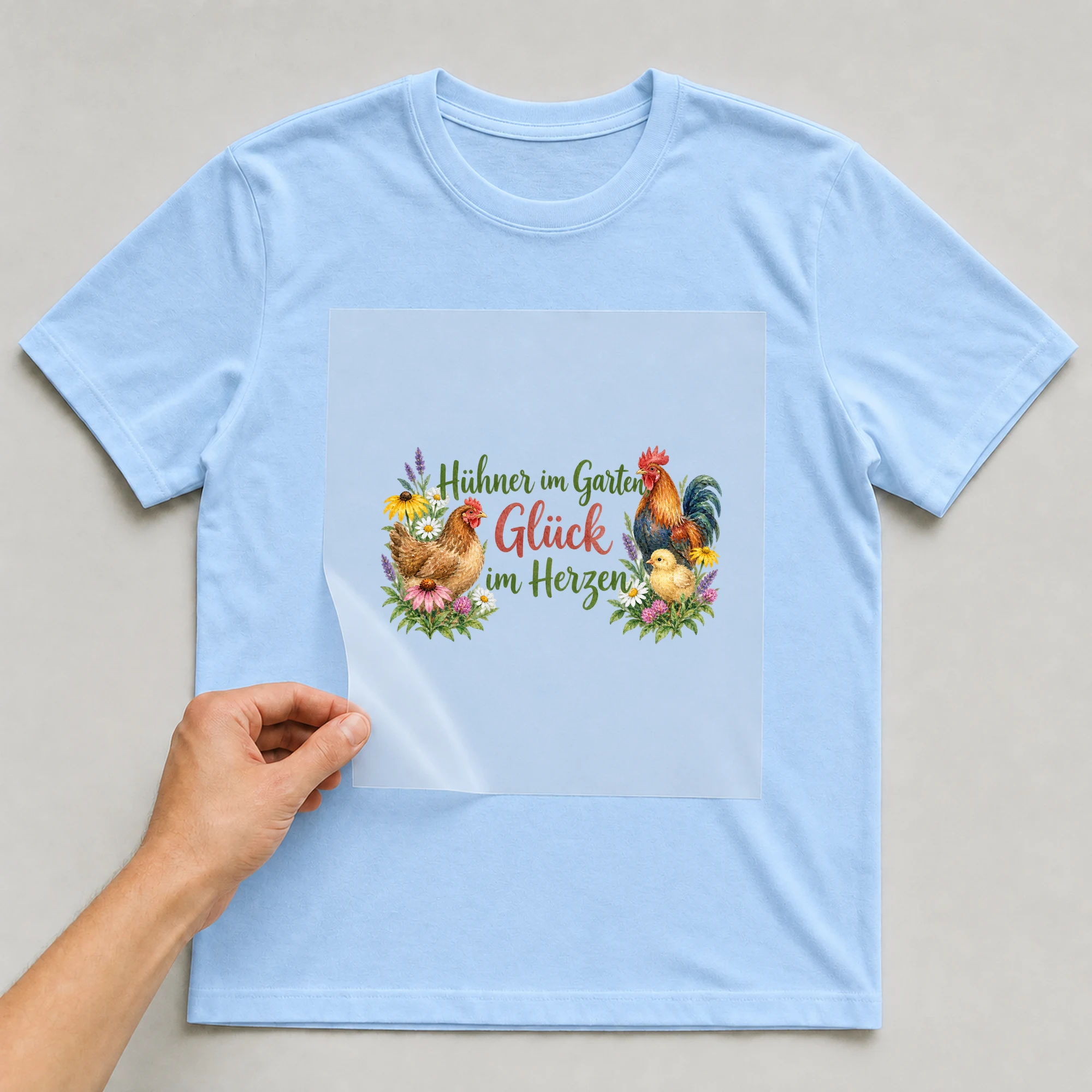 Bügelbild „Hühner im Garten, Glück im Herzen“ auf babyblauem T-Shirt, Folie wird aufgelegt