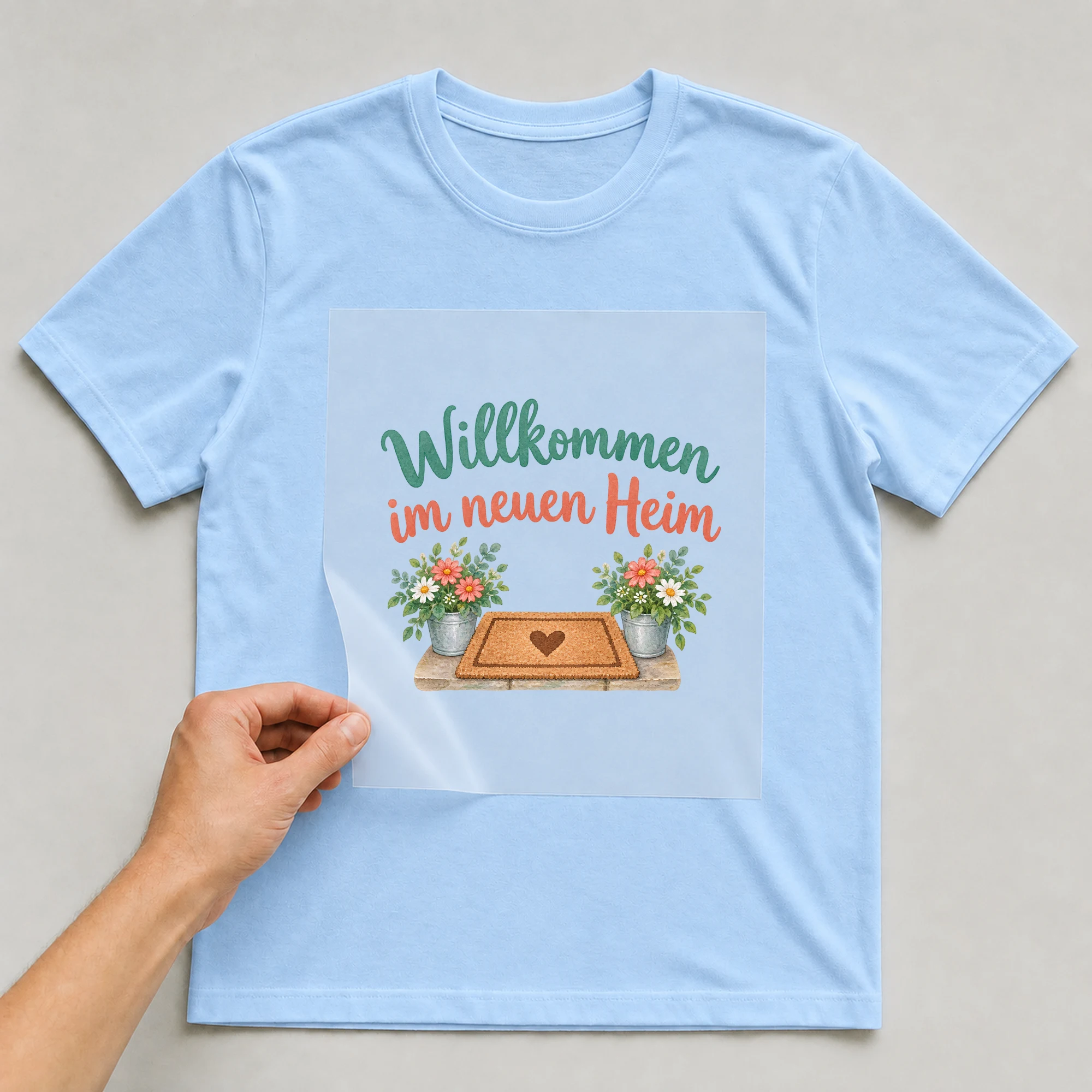 Bügelbild „Willkommen im neuen Heim“ auf babyblauem T-Shirt, Folie wird aufgelegt