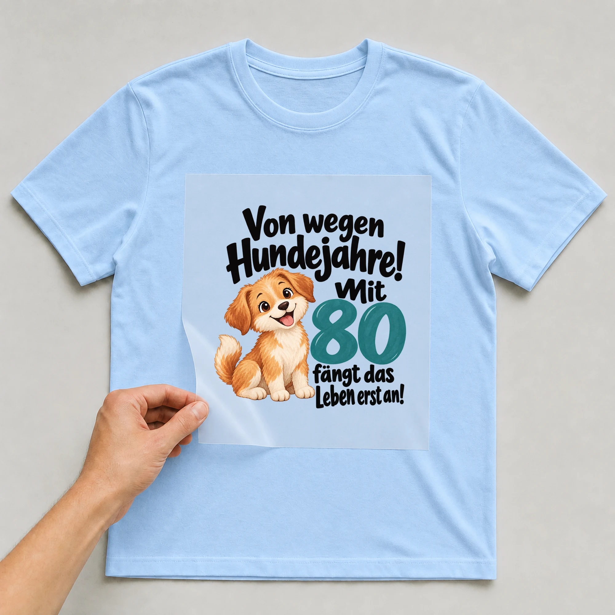 Bügelbild „Von wegen Hundejahre! Mit 80 fängt das Leben erst an!“ auf babyblauem T-Shirt, Folie wird aufgelegt