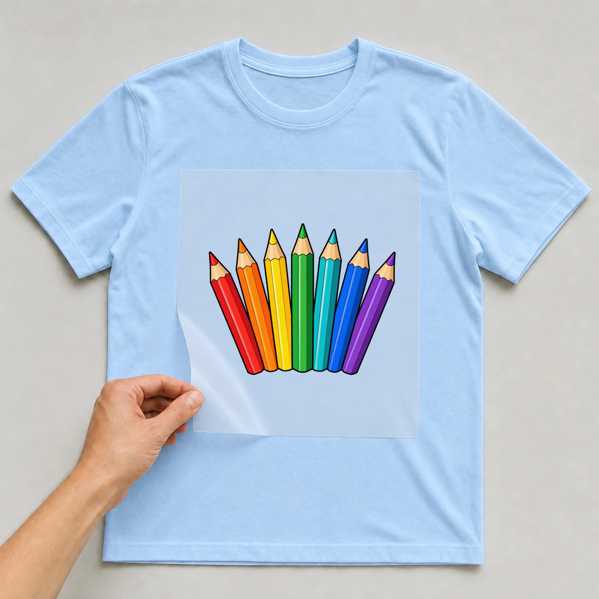 Bügelbild „Buntstifte“ auf babyblauem T-Shirt, Folie wird aufgelegt